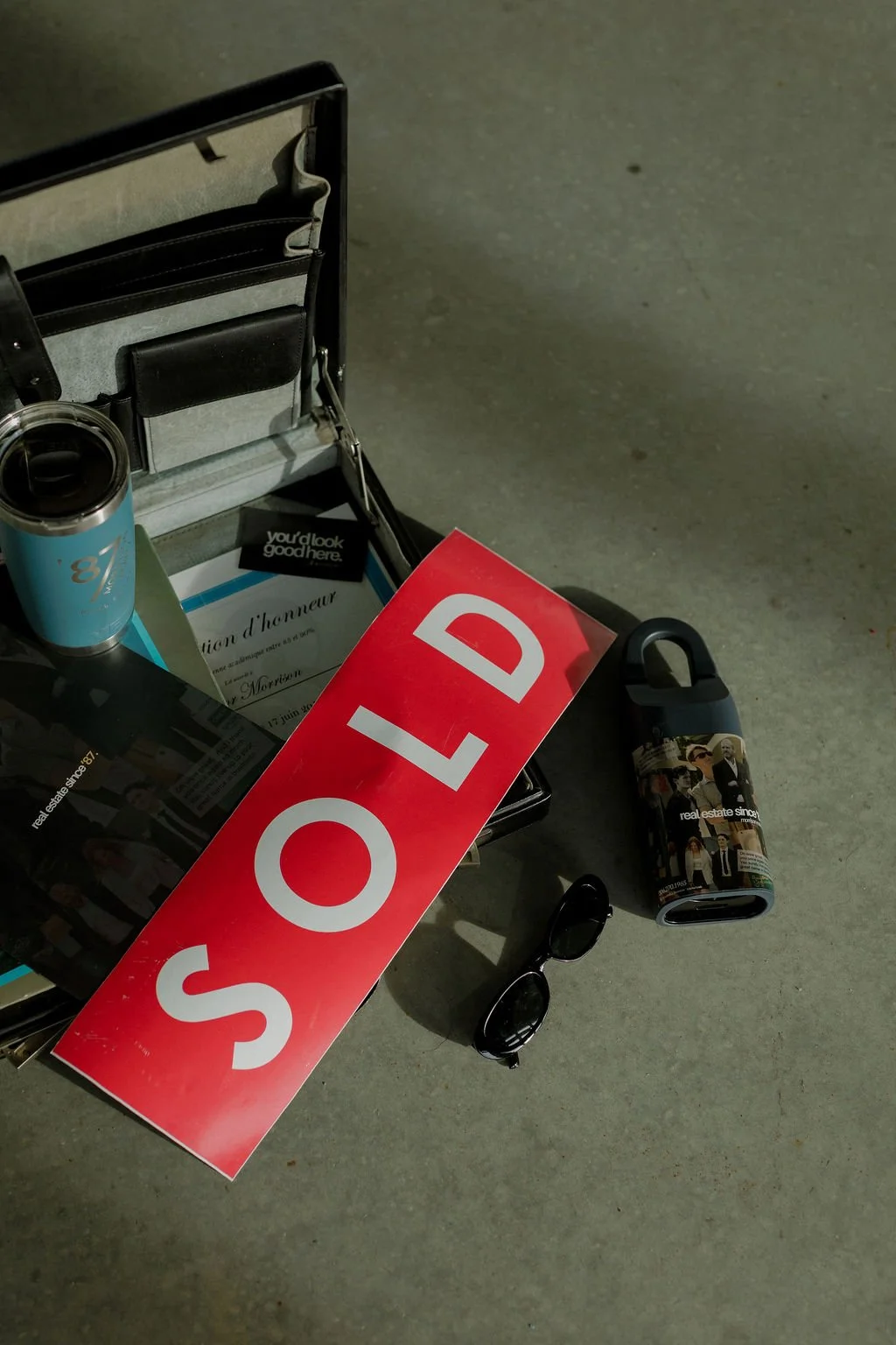 A small table with a red and white 'SOLD' sign, sunglasses, a tumbler with Morrison Realty '87 branded photos, a brochure, and a black card, on a concrete floor.