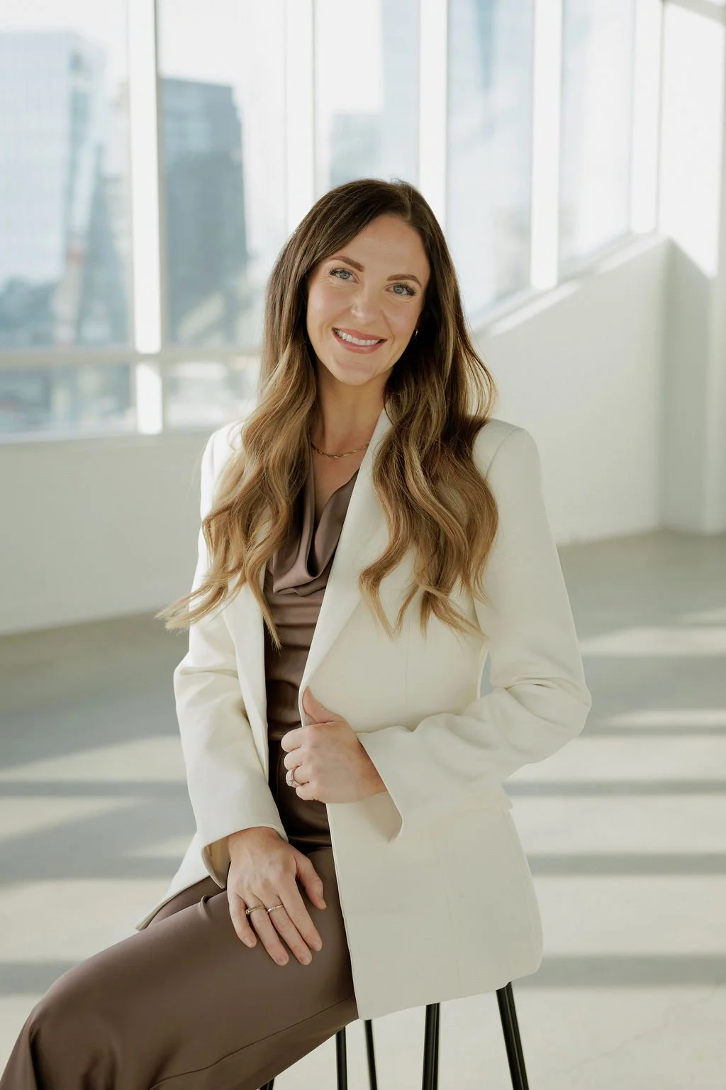 A woman with long wavy brown hair smiles while sitting on a stool in a bright room with large windows and city skyline in the background. She is wearing a beige blazer over a brown blouse.