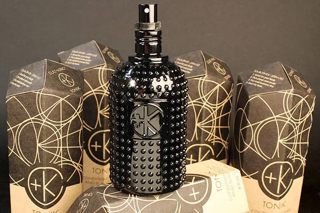 A black perfume or cologne spray bottle with decorative black studs, surrounded by cardboard boxes with geometric patterns, against a plain background.