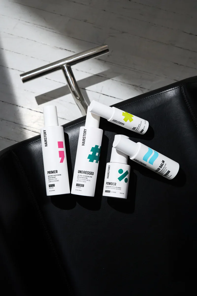 Collection of HAIRSTORY hair styling products including primer, undressed detangling spray, powder, and balm, placed on a black leather surface with a metal T-shaped handle in the background.