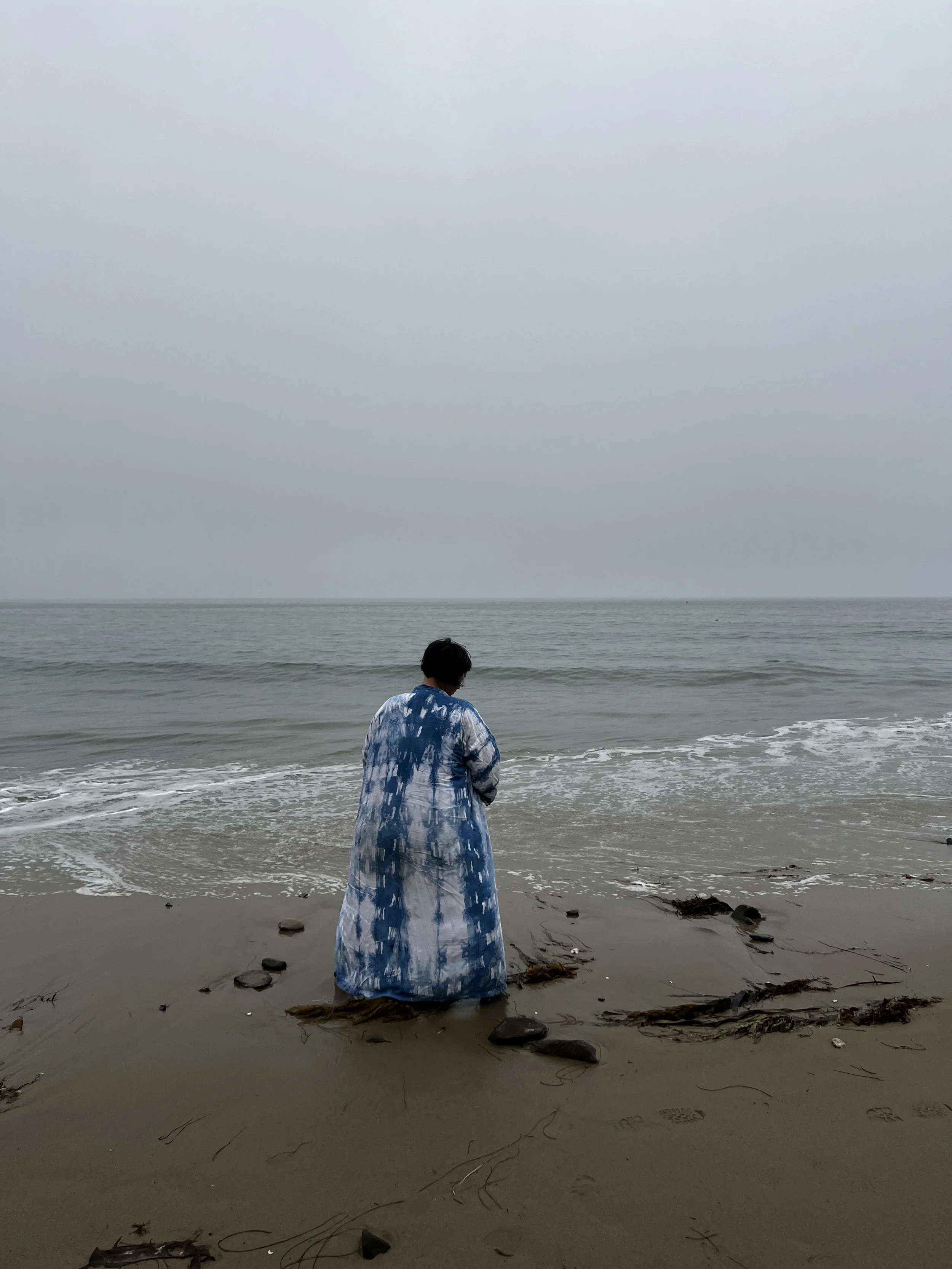 A person wearing a blue and white tie-dye robe standing on a beach, facing the ocean under an overcast sky.