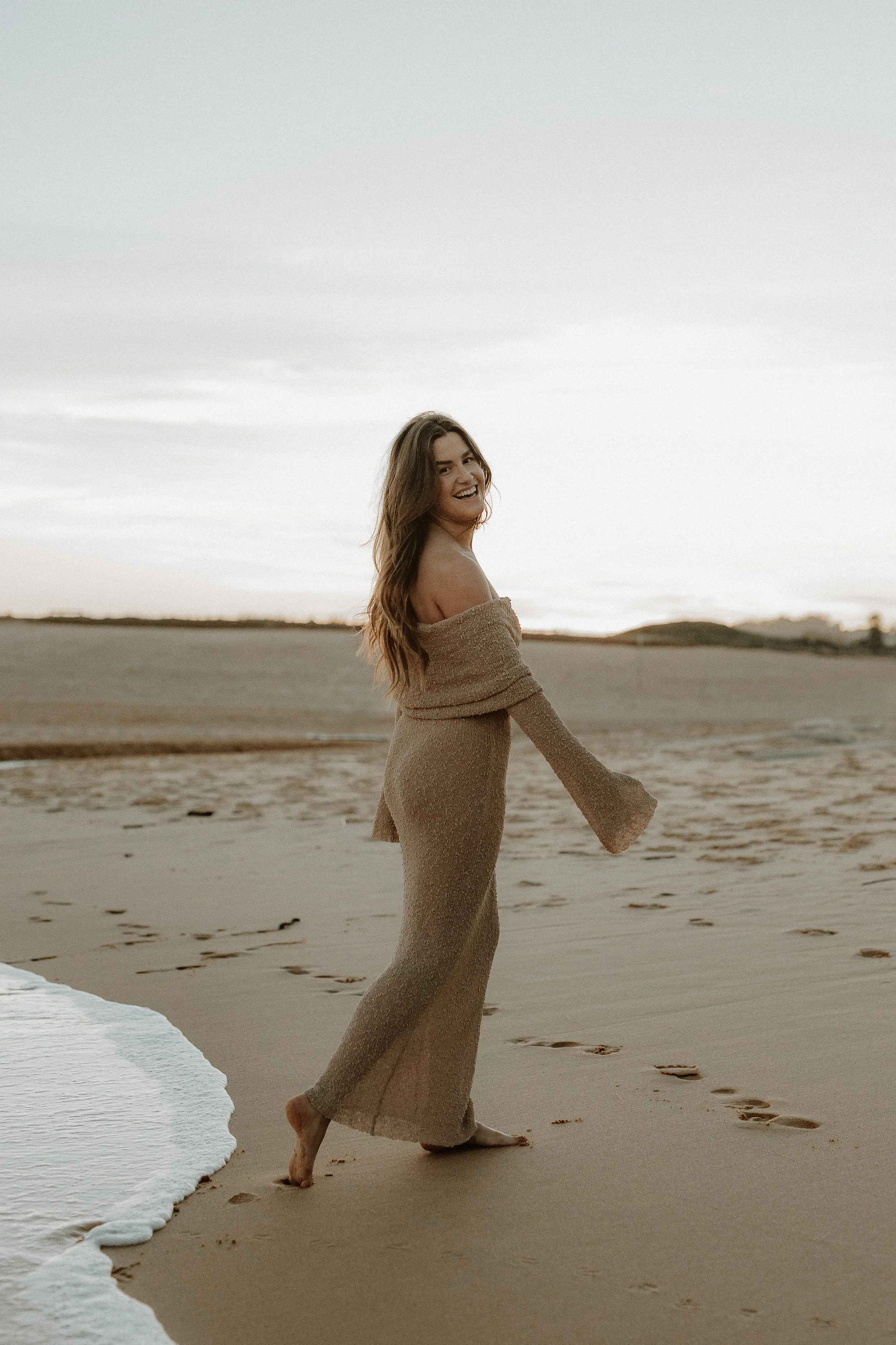 A woman in a beige dress walking barefoot on the beach near the shoreline, with ocean waves and distant hills visible in the background at sunset.