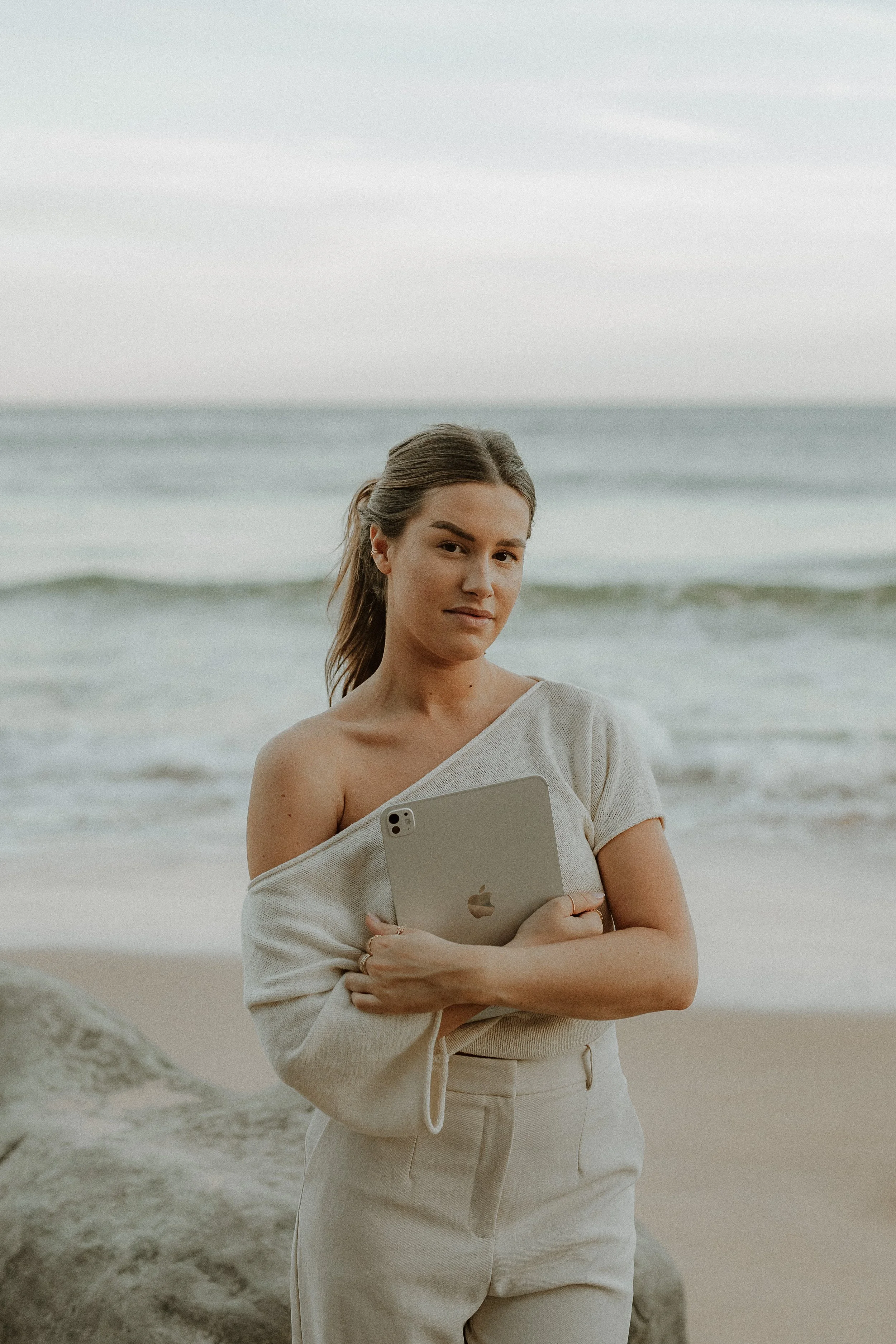 A young woman standing on a beach holding an Apple iPad, with the ocean and sky in the background.