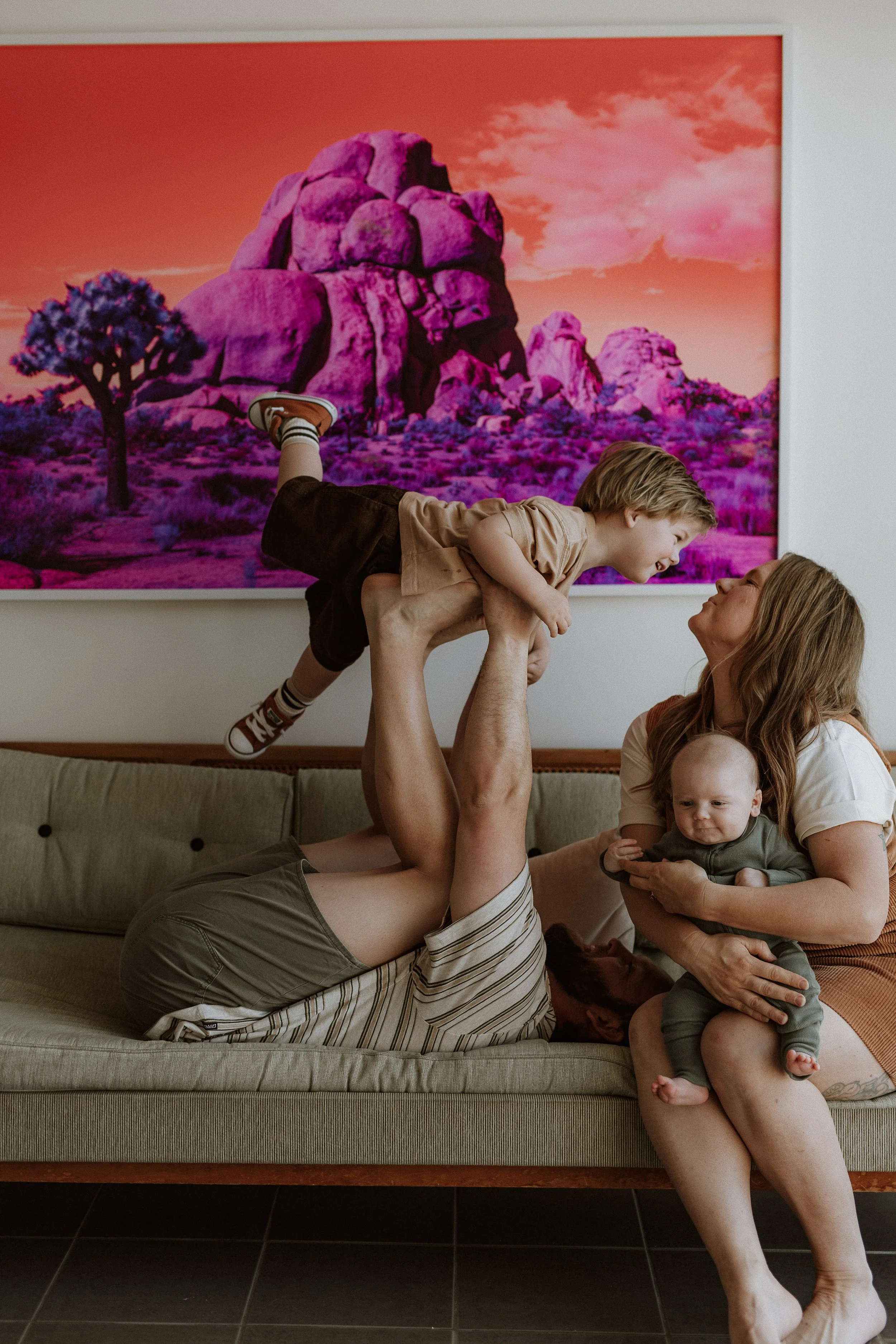 A family is playing on a beige sofa in a room with dark tile flooring. An adult is lying on their back, holding up a young boy in a brown shirt and shorts, who appears to be smiling and playing. The woman next to them is holding another young child i