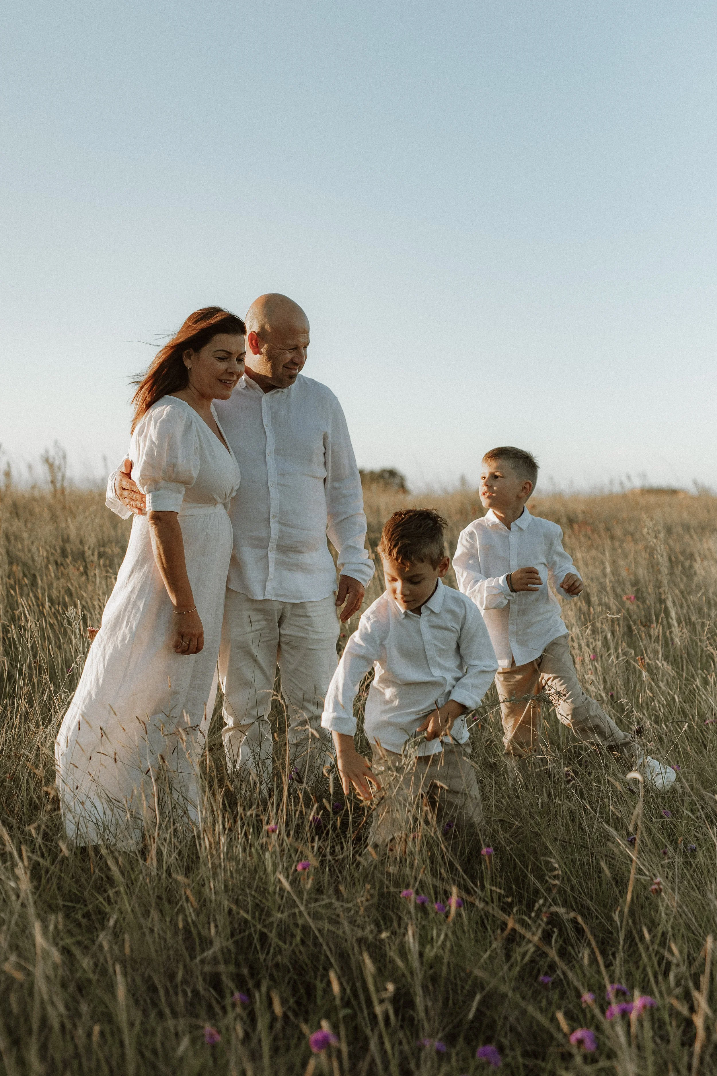 A family of five, dressed in white, walking through a grassy field during sunset. The family includes a woman, a bald man, and three young boys. The woman and man are holding hands, and the boys are playing and exploring in the grass.