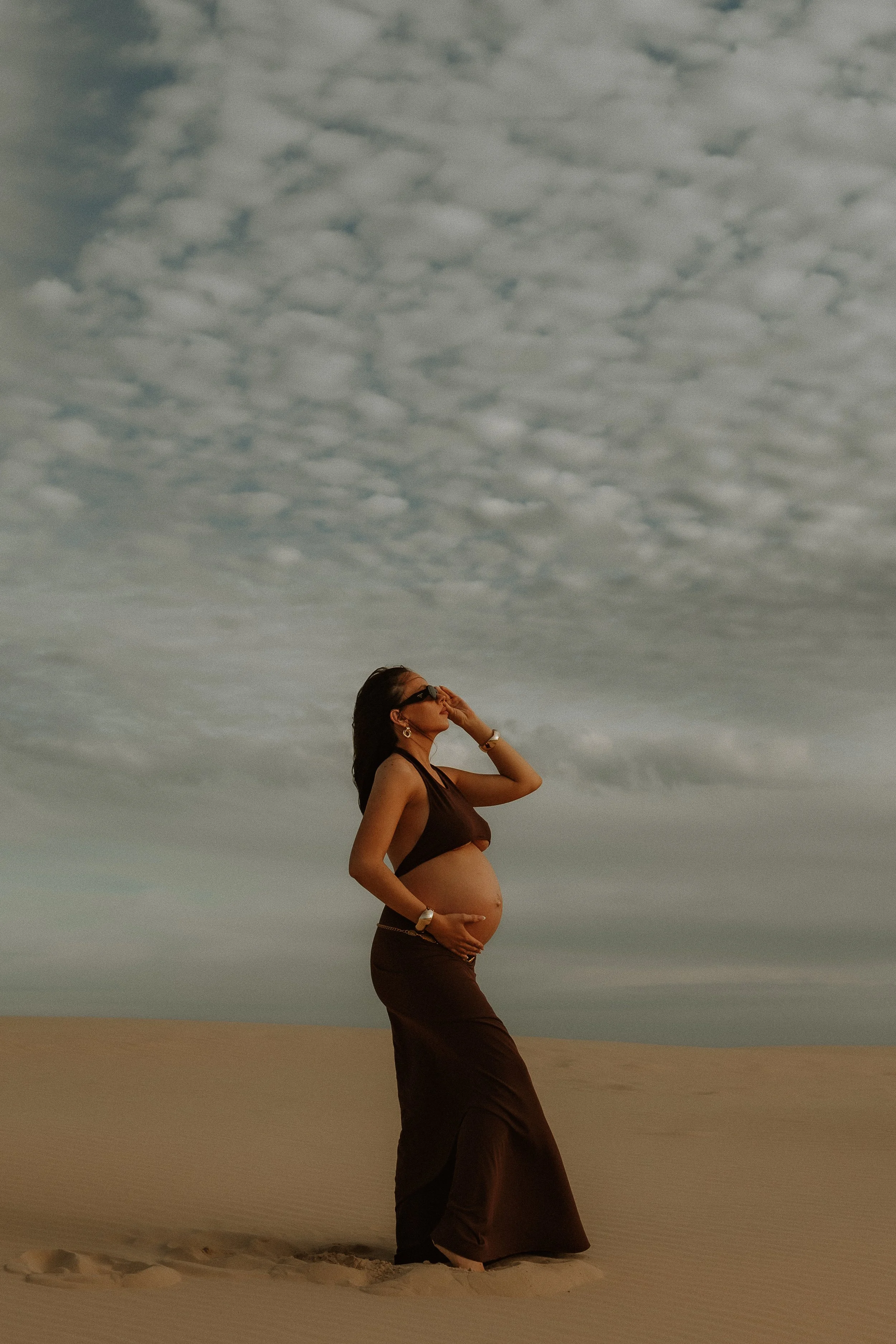 A pregnant woman standing on a sandy beach, wearing a dark crop top and long skirt, touching her belly while looking at the cloudy sky above.