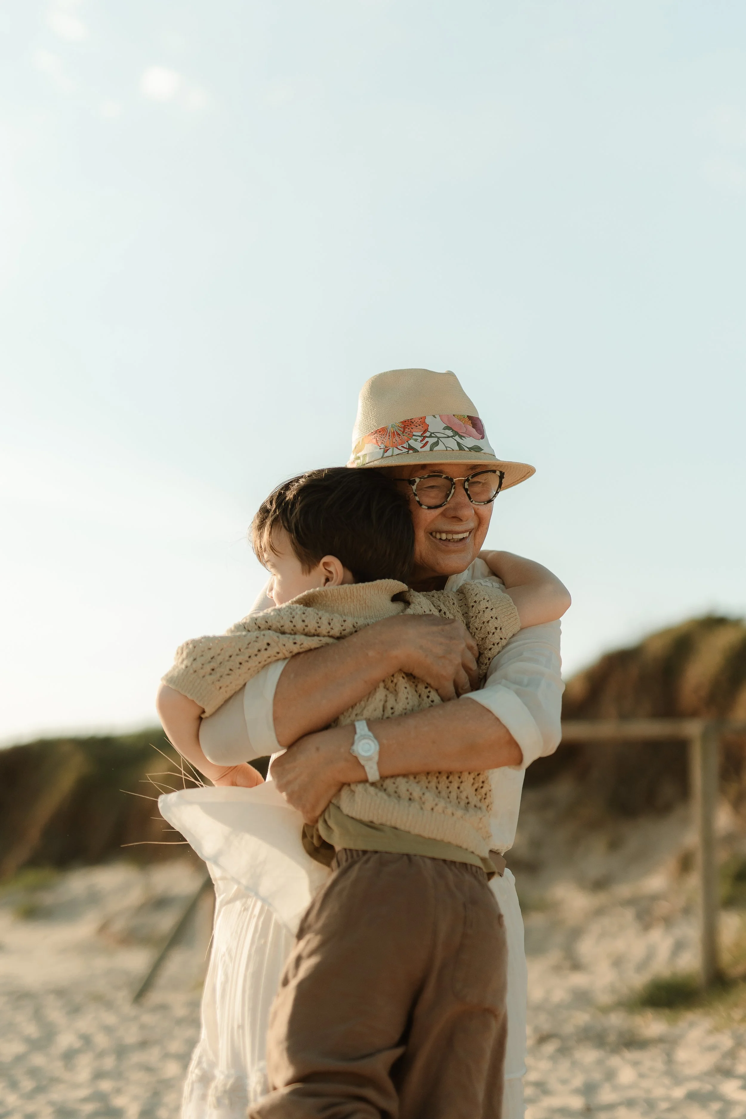 A woman wearing glasses and a beige hat with a floral band is hugging a young boy, both smiling, outdoors on a sunny day.