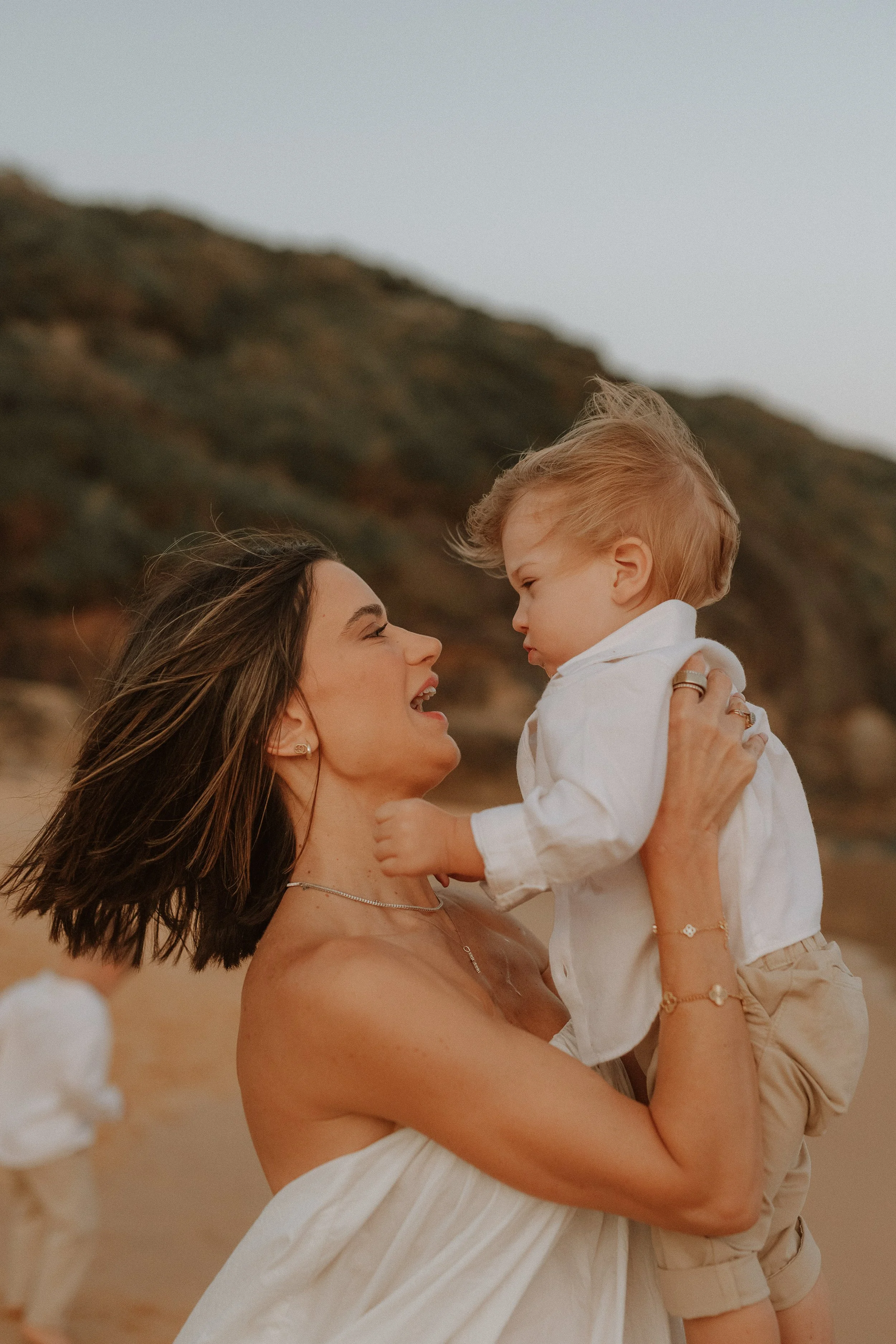 A woman with long brown hair holding a young boy with blond hair at the beach during sunset, with a rocky hill in the background.