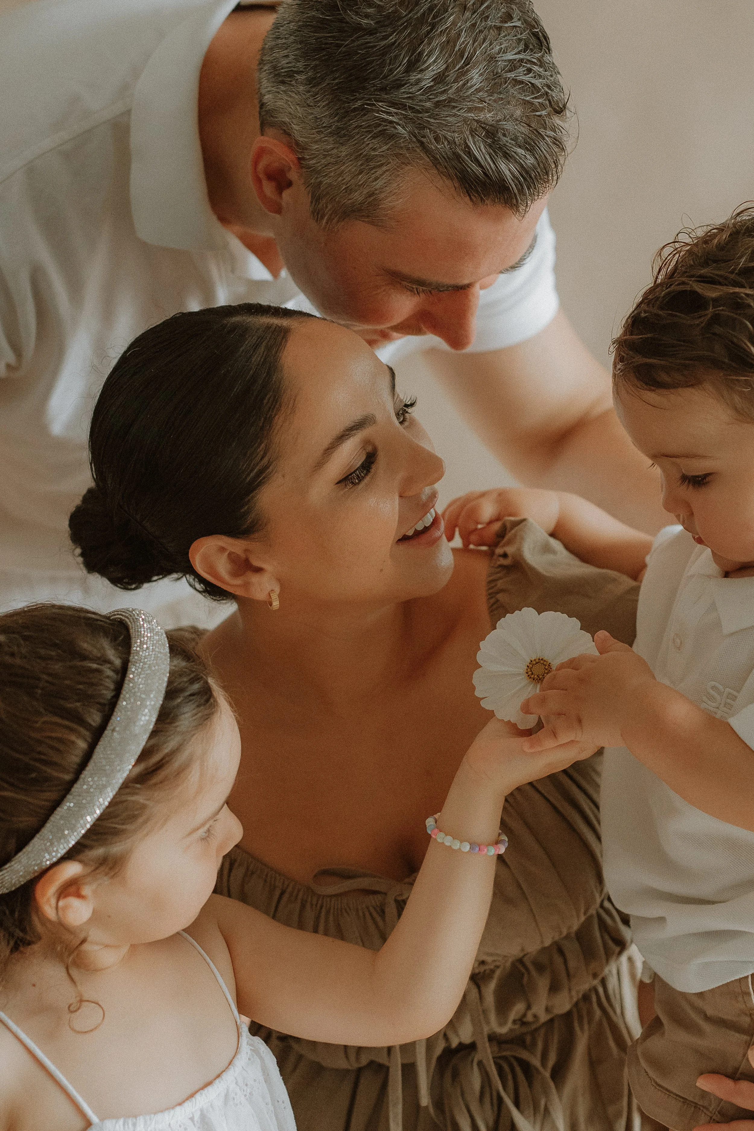 A family of four with a mother, father, and two young children, sharing a close moment. The mother is smiling while holding a white flower, with the children and father gathered around her in an intimate setting.