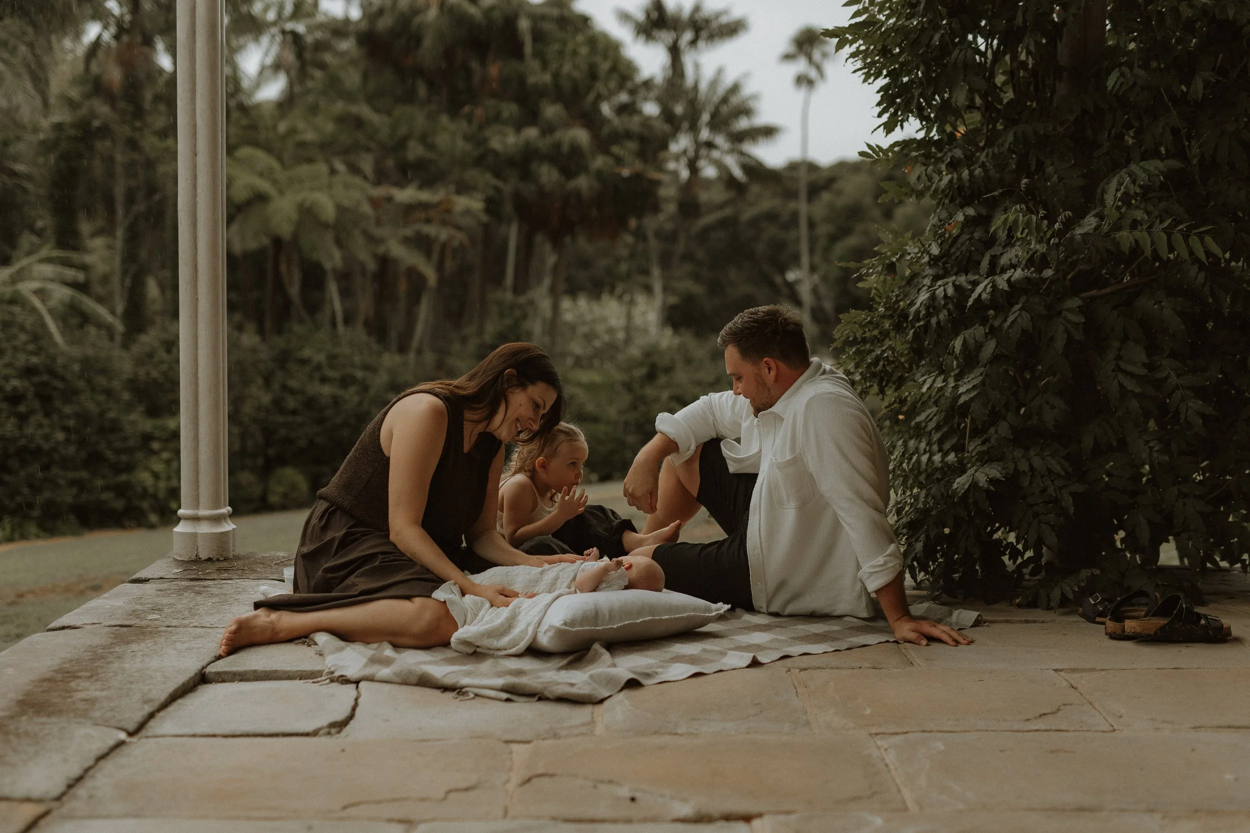 A family sitting on a blanket outdoors near a lake, including two adults, a child, and a baby, with trees and a cloudy sky in the background.