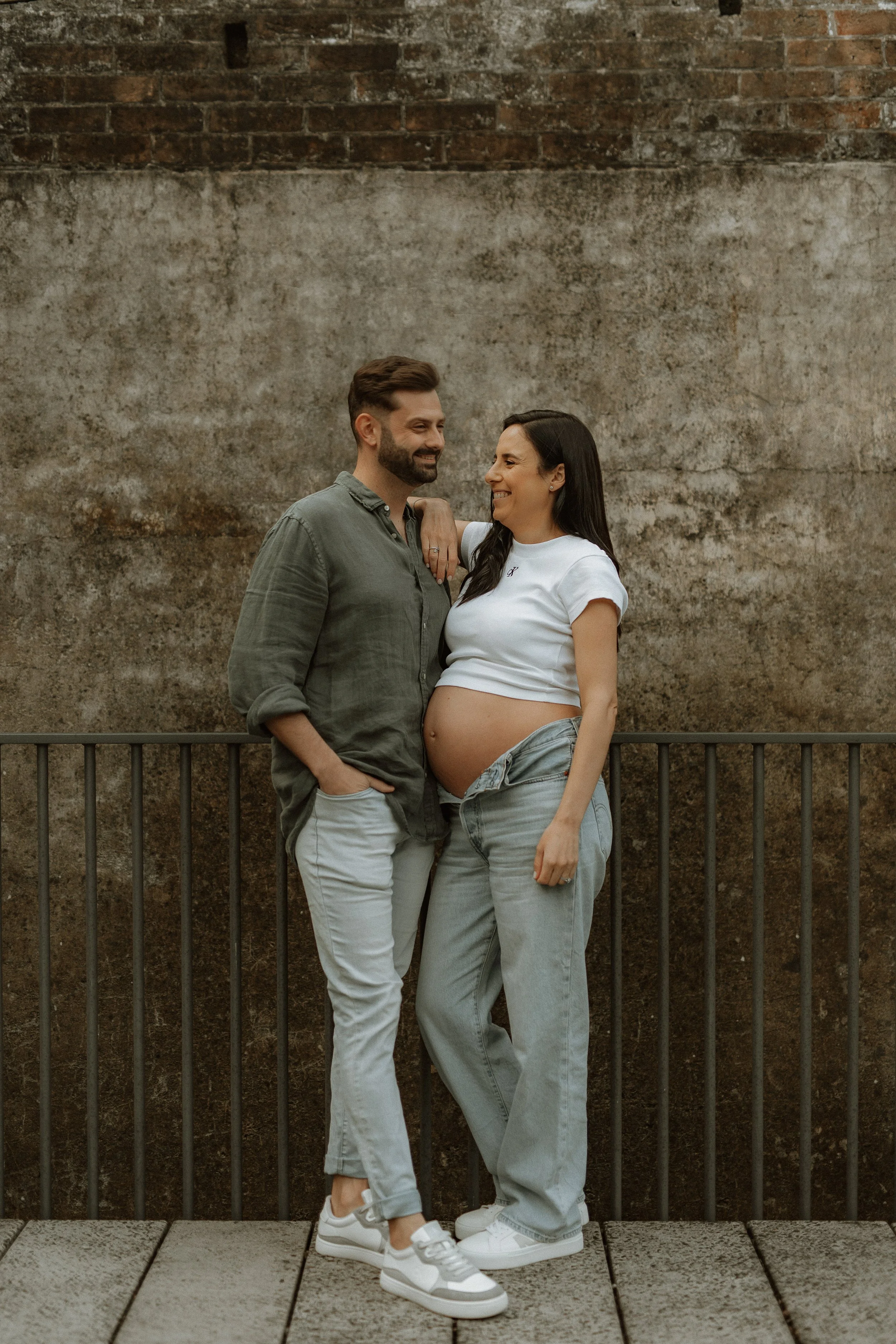 A couple stands close together against a textured brick wall. The woman is pregnant and wearing a white t-shirt and gray jeans, smiling and looking at the man. The man is wearing a green button-up shirt and light-colored pants, smiling and looking at