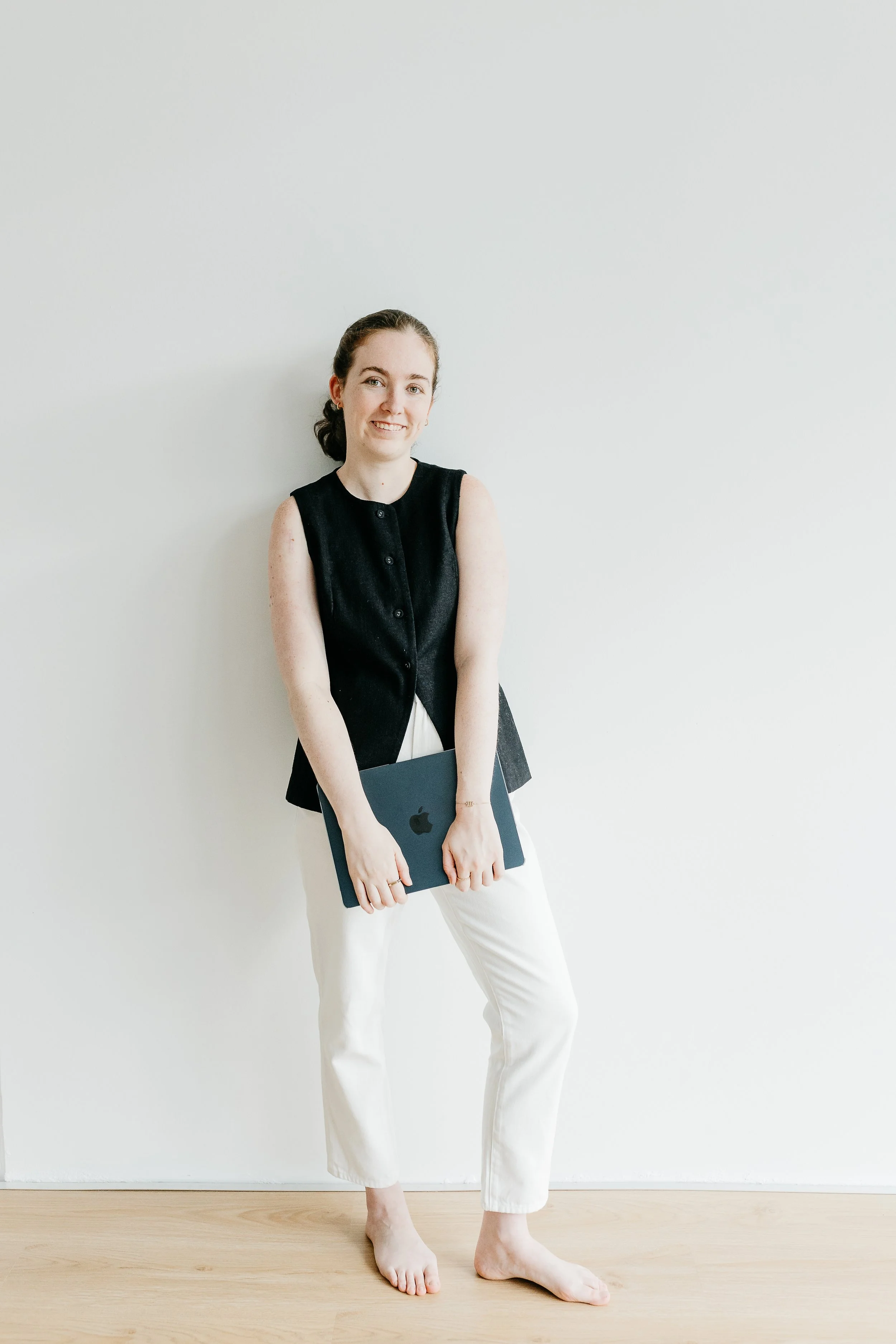 A woman standing barefoot against a plain white wall, holding a closed MacBook laptop, dressed in a black sleeveless top and white pants, smiling softly.