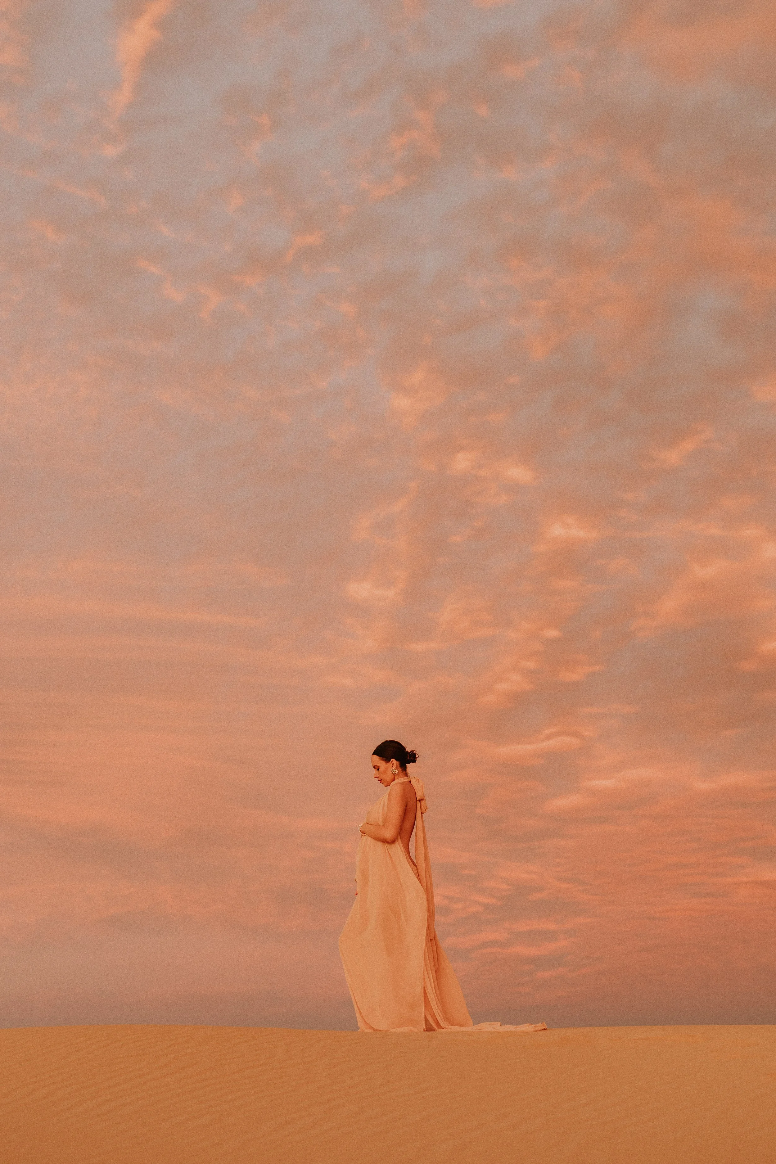 A woman in a flowing beige dress stands alone in a desert landscape at sunset, with a sky filled with pink and orange clouds.
