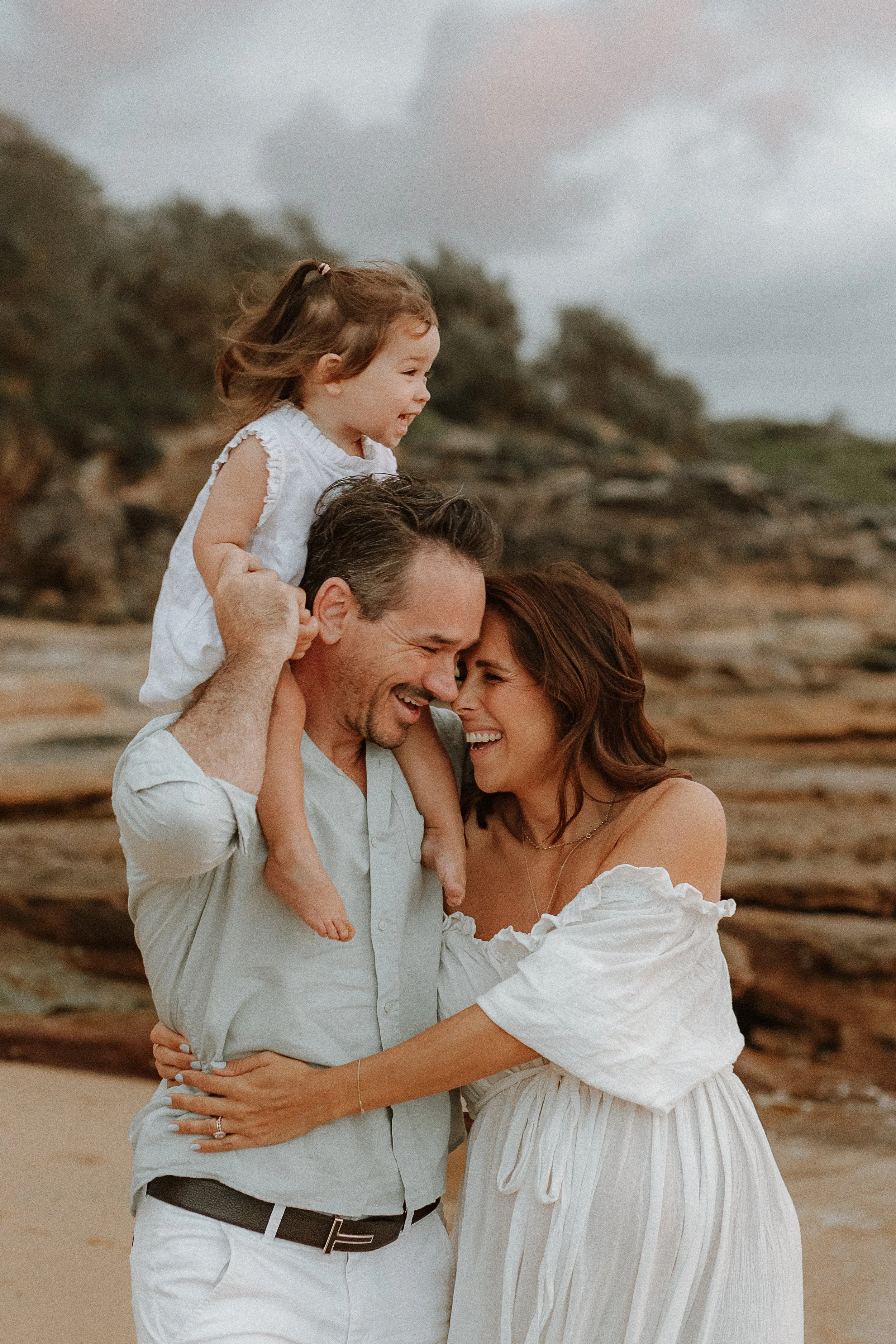 A happy family of three at the beach, parents holding their young daughter on their shoulders, all smiling and enjoying each other's company on a cloudy day.