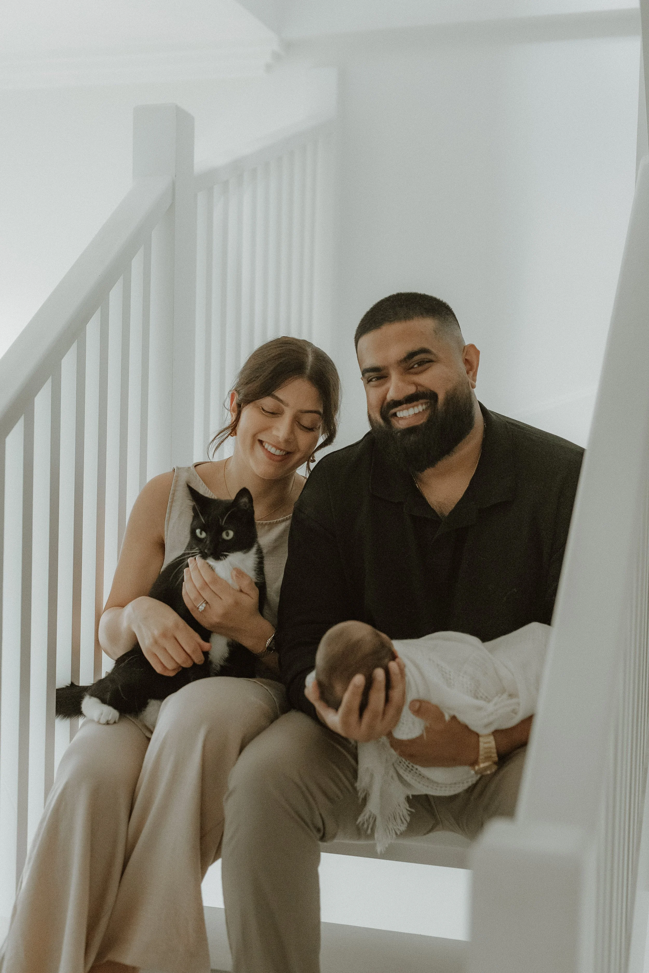A family sitting on the stairs, smiling, with a woman holding a black and white cat and a man holding a newborn baby.