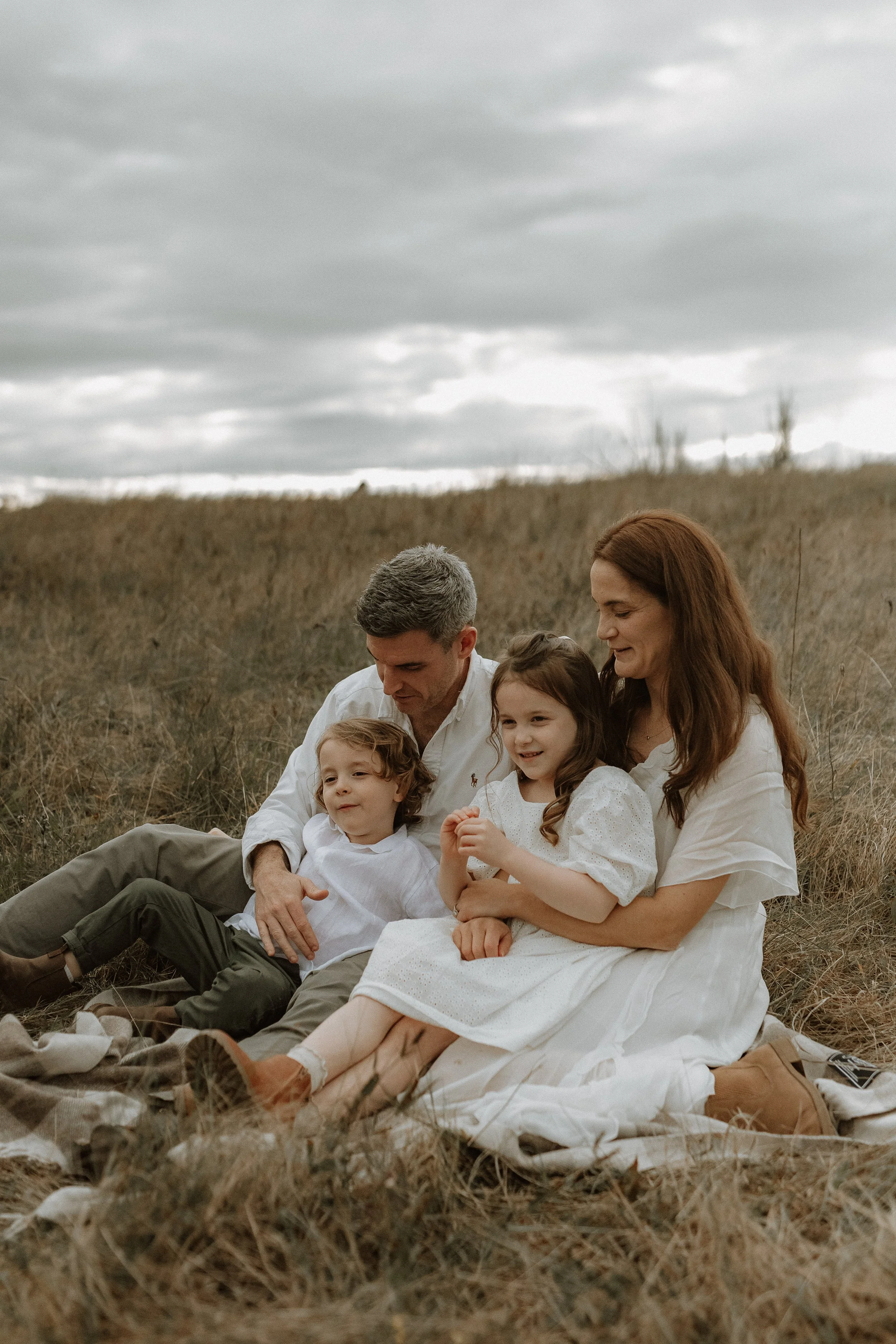 A family of six sitting together outdoors in a grassy field under a cloudy sky. The family includes two adults and four children, all dressed in light-colored clothing, smiling and relaxing.