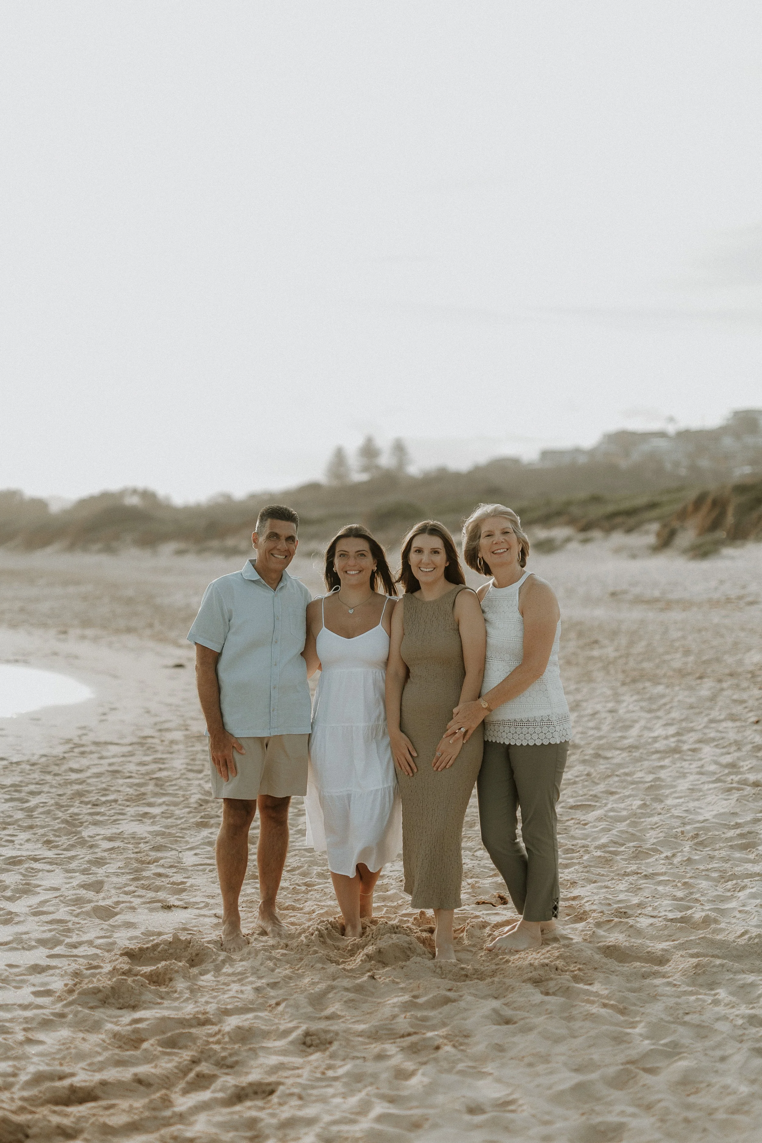 A group of five people standing on a sandy beach with houses and trees in the background. They are smiling and dressed in casual summer clothing.