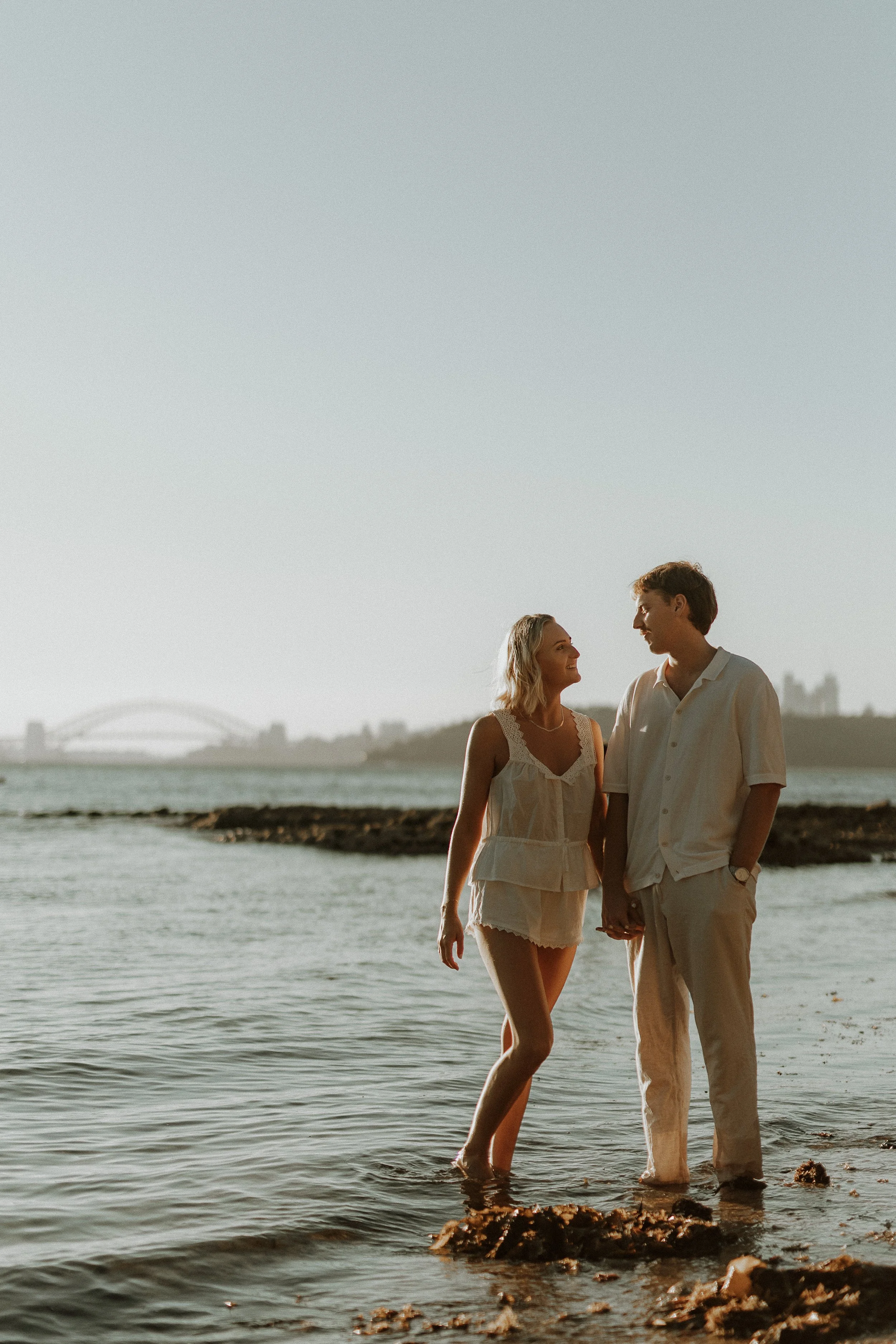 A young couple holding hands and smiling at each other while walking along the shoreline at sunset, with a city bridge visible in the background.