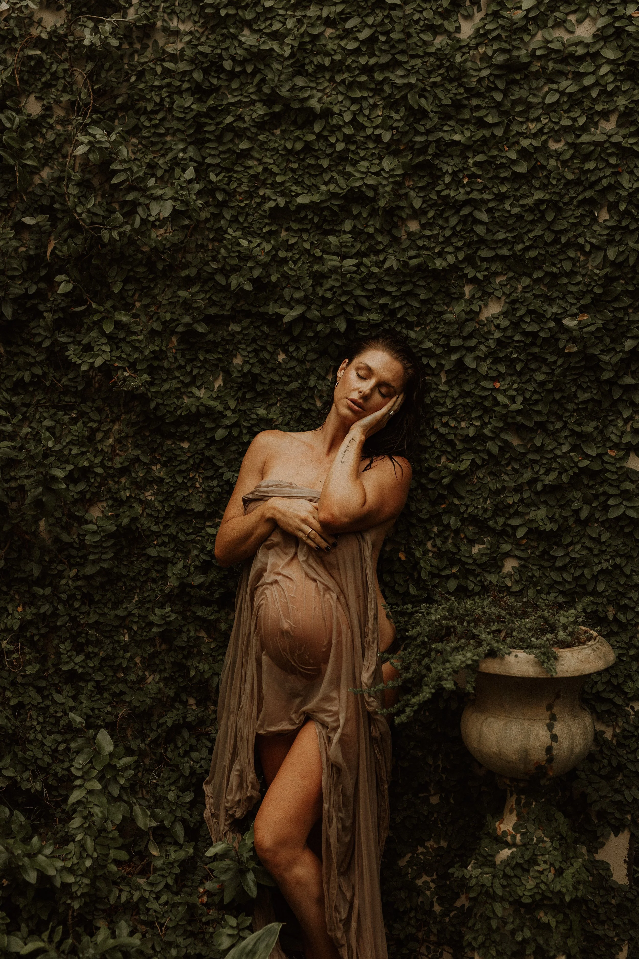 A woman with wet hair and tan clothing poses against a dense green ivy-covered wall, with one knee on the ground and her eyes closed, touching her face with one hand.