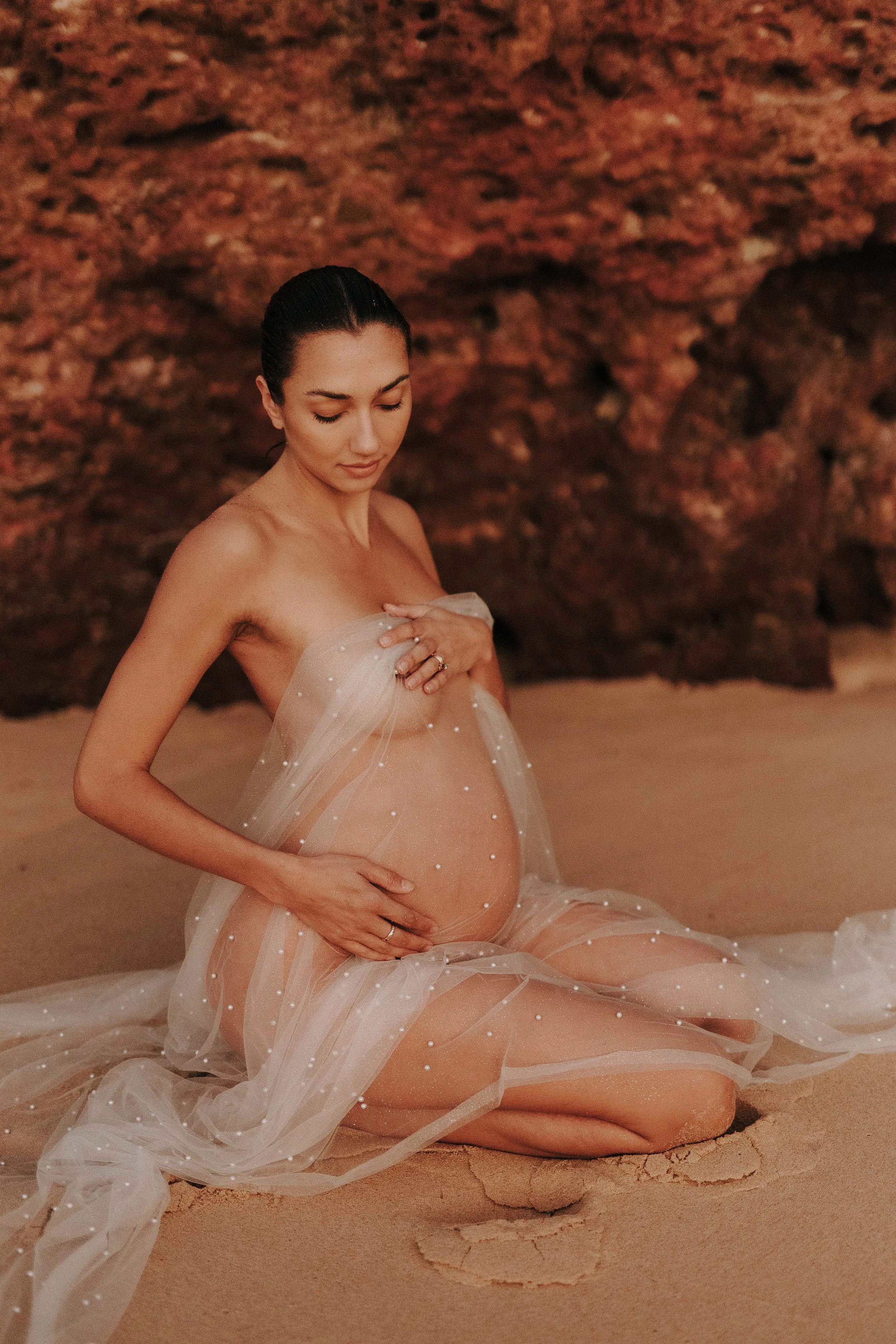 A pregnant woman with dark hair styled back, sitting on the sandy ground with a rocky background, draped in a sheer, light-colored fabric with small pearl-like accents, gently touching her belly and looking down