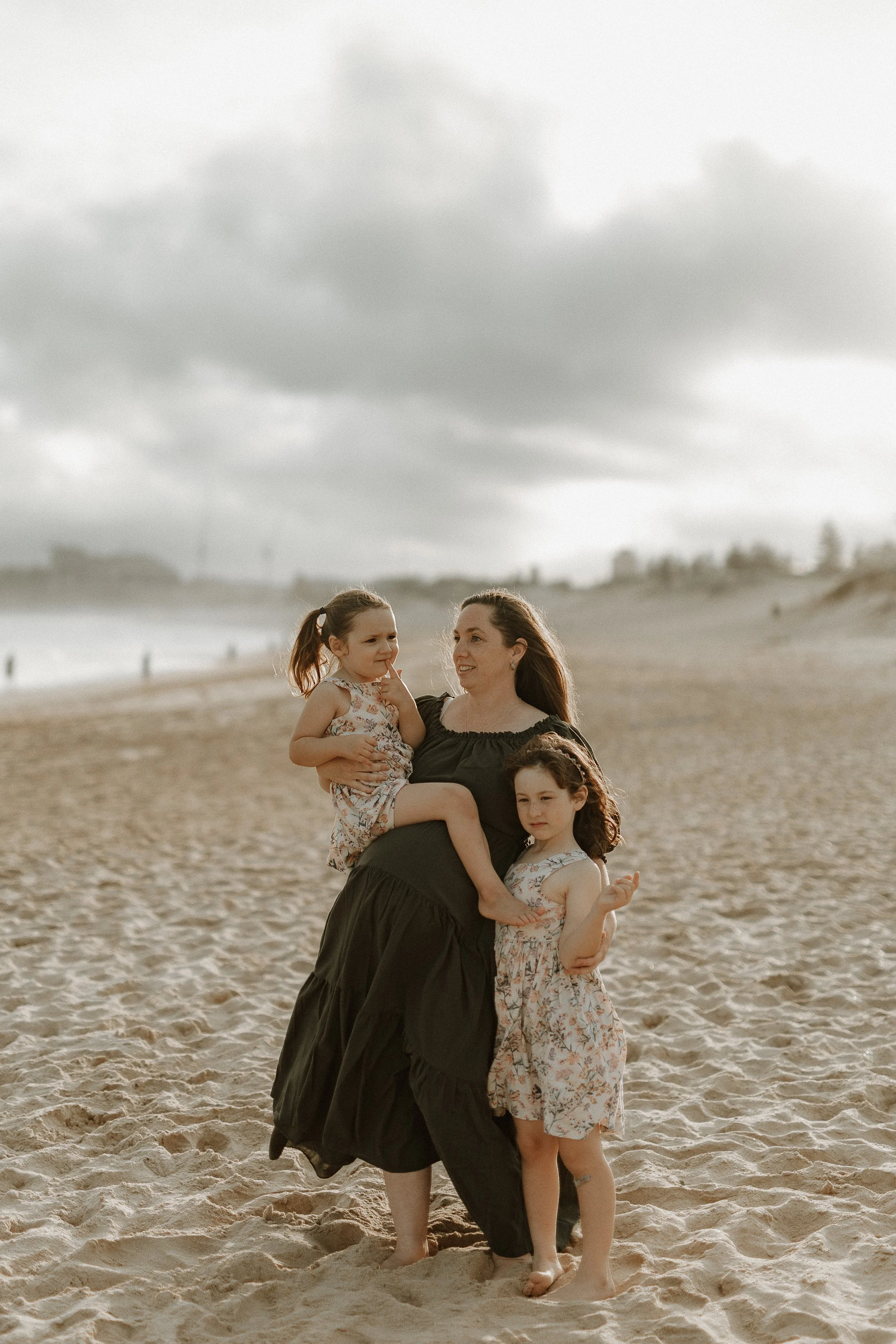 Woman with two young girls on a beach, cloudy sky, sandy background.