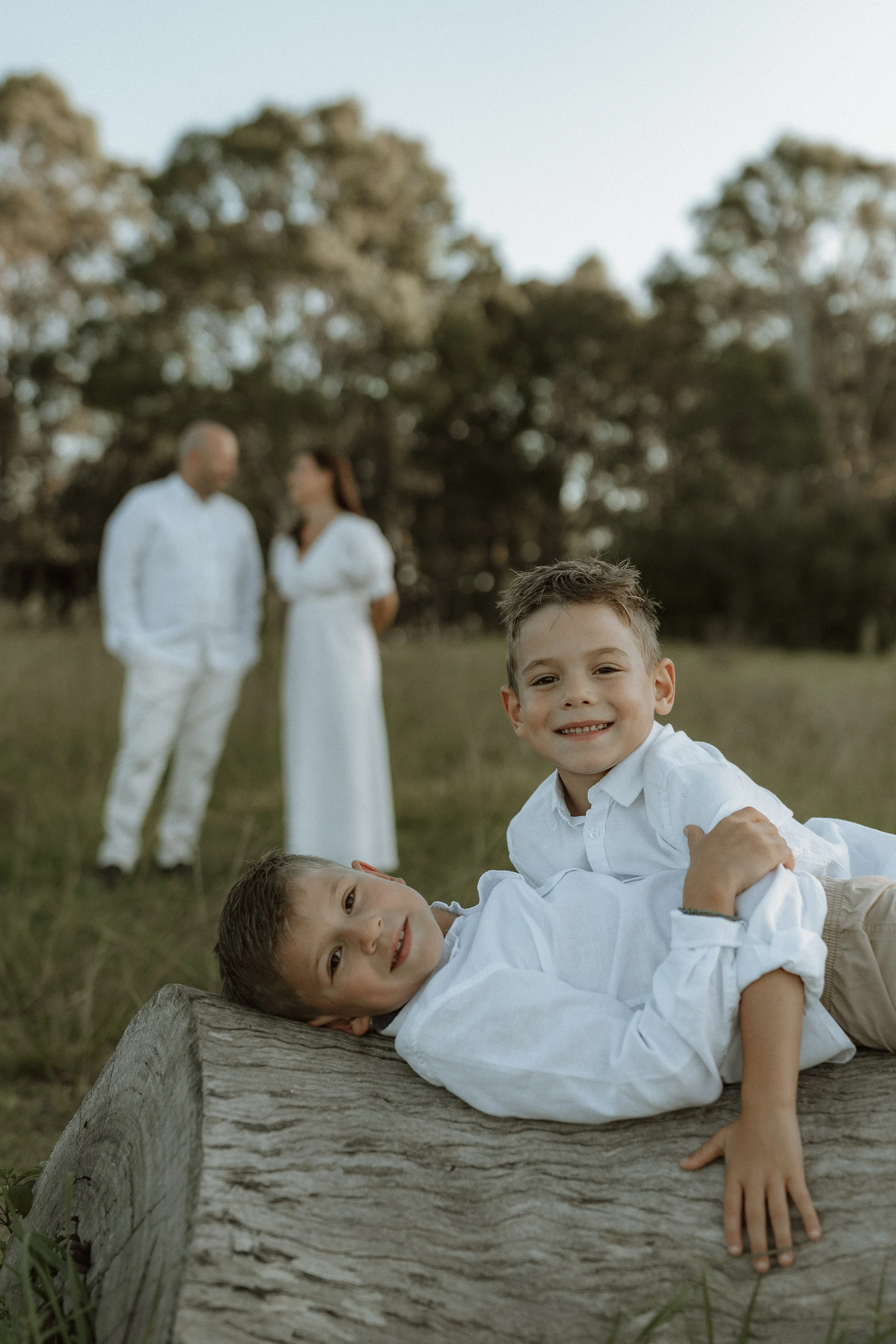Two smiling boys in white shirts are playing on a log in a grassy field with trees in the background while a man and a woman, also dressed in white, stand and talk in the distance.