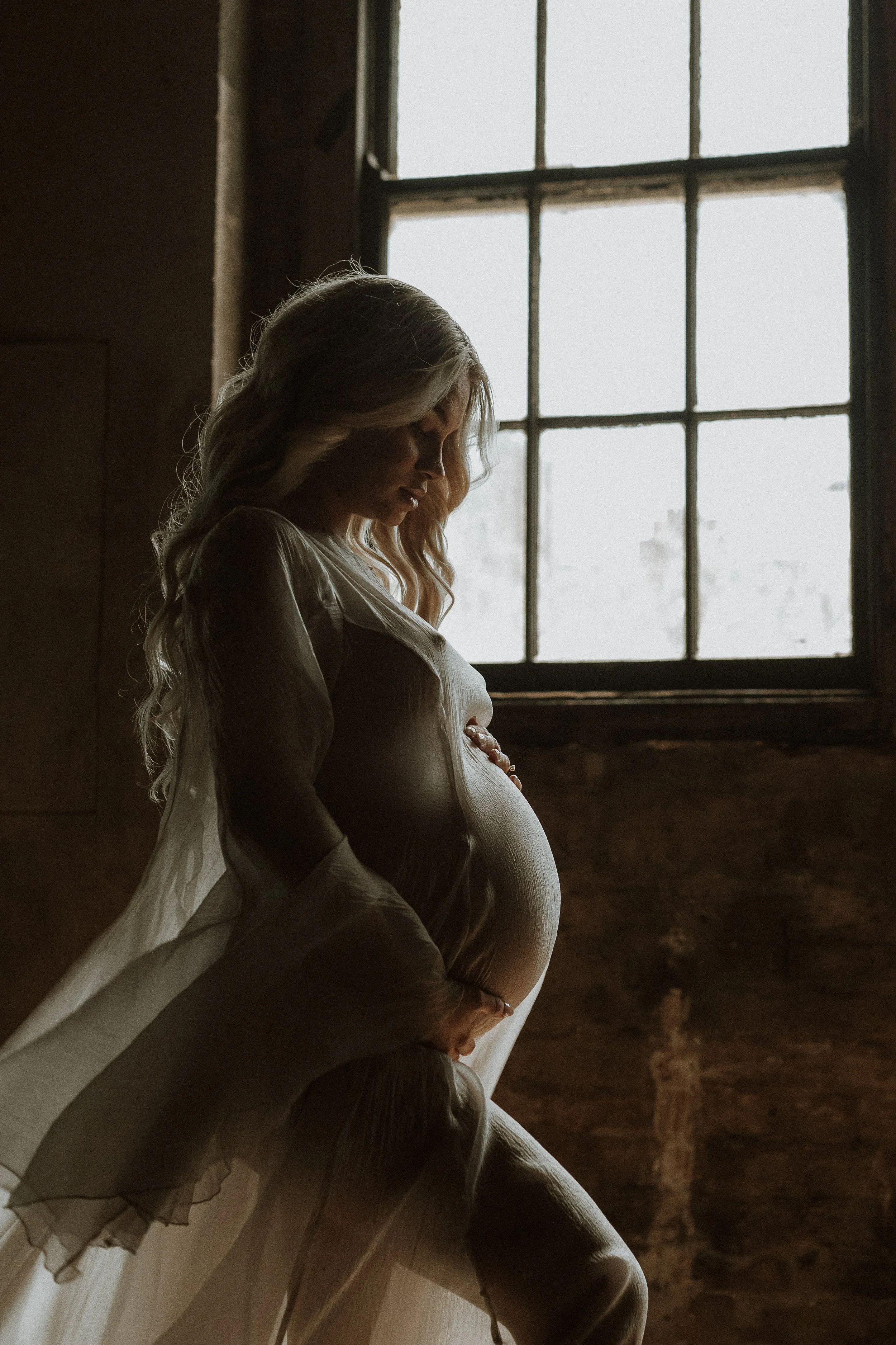A pregnant woman with long blonde hair sitting by a window, gazing downward, in a dimly lit room.