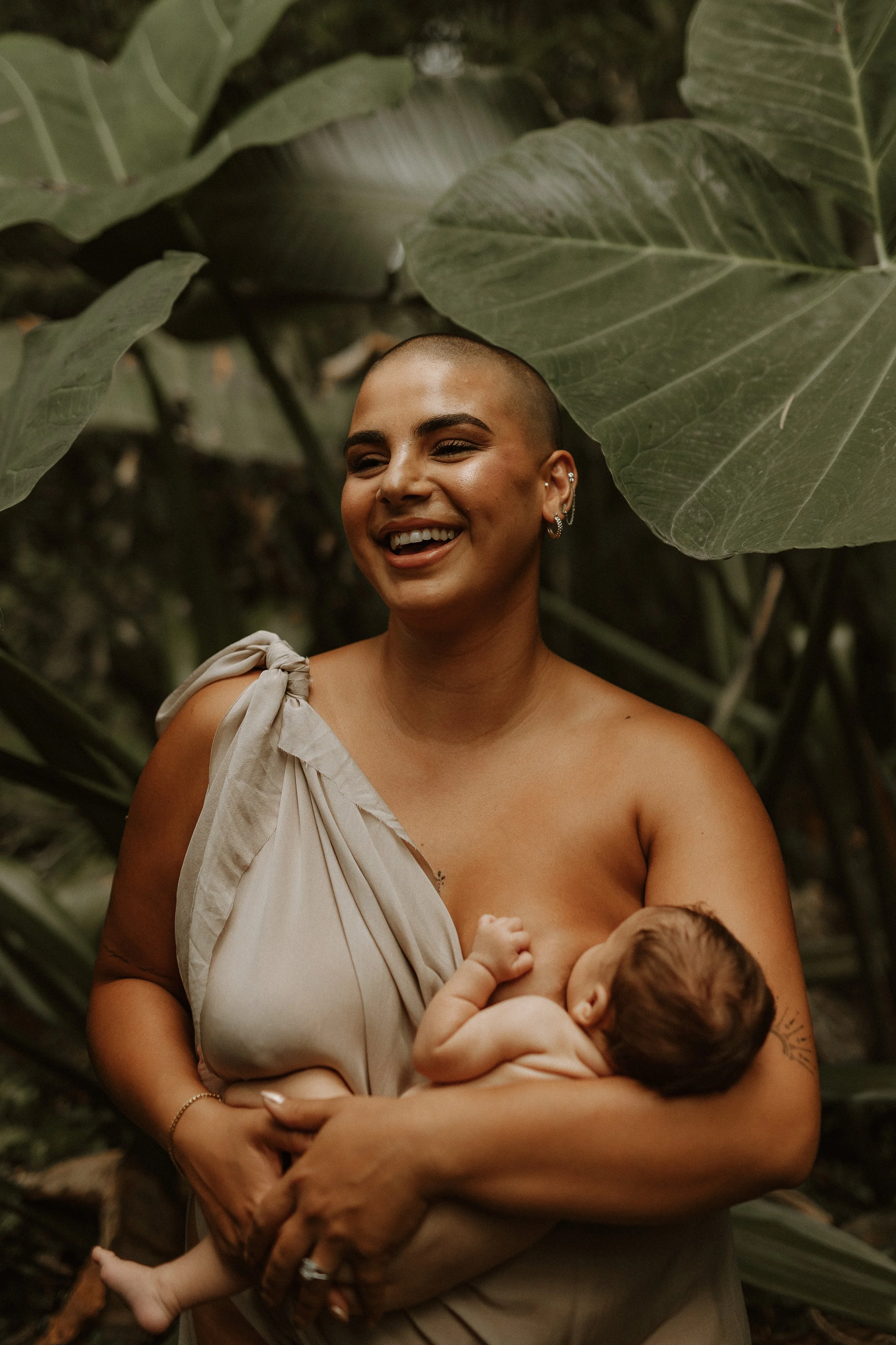 A woman with a shaved head smiling while breastfeeding a baby in an outdoor jungle setting with large green leaves.