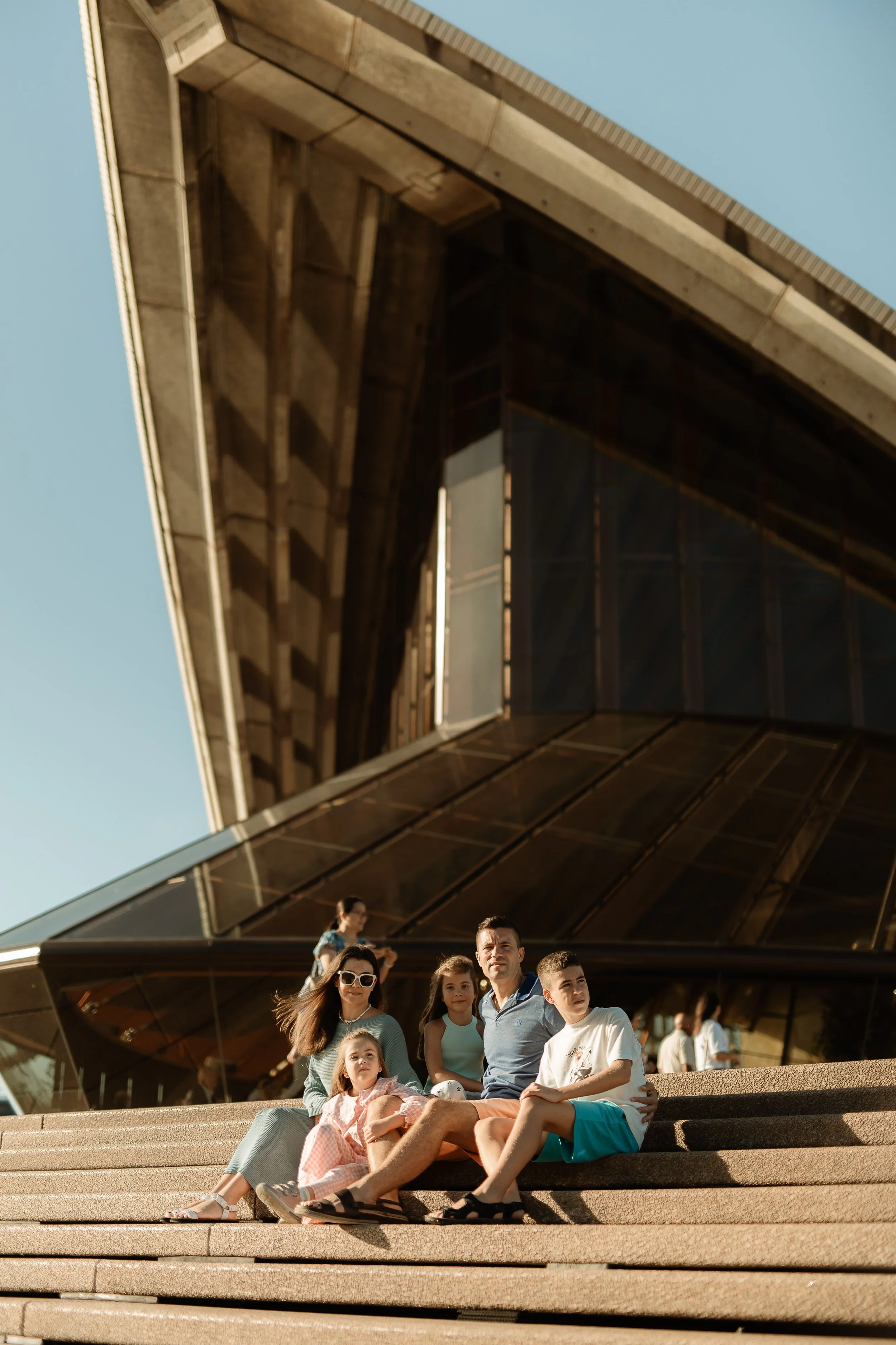 Family sitting on steps outside a modern building with angular architectural design and large glass windows.