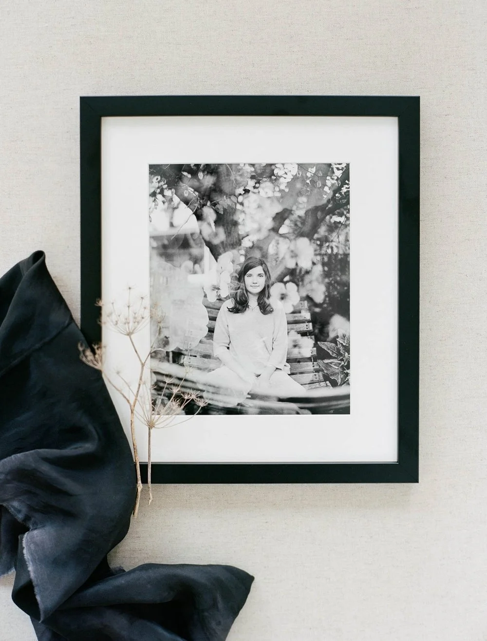 Framed black and white photograph of a person sitting, surrounded by flowers, with a black fabric and dried branch beside the frame.