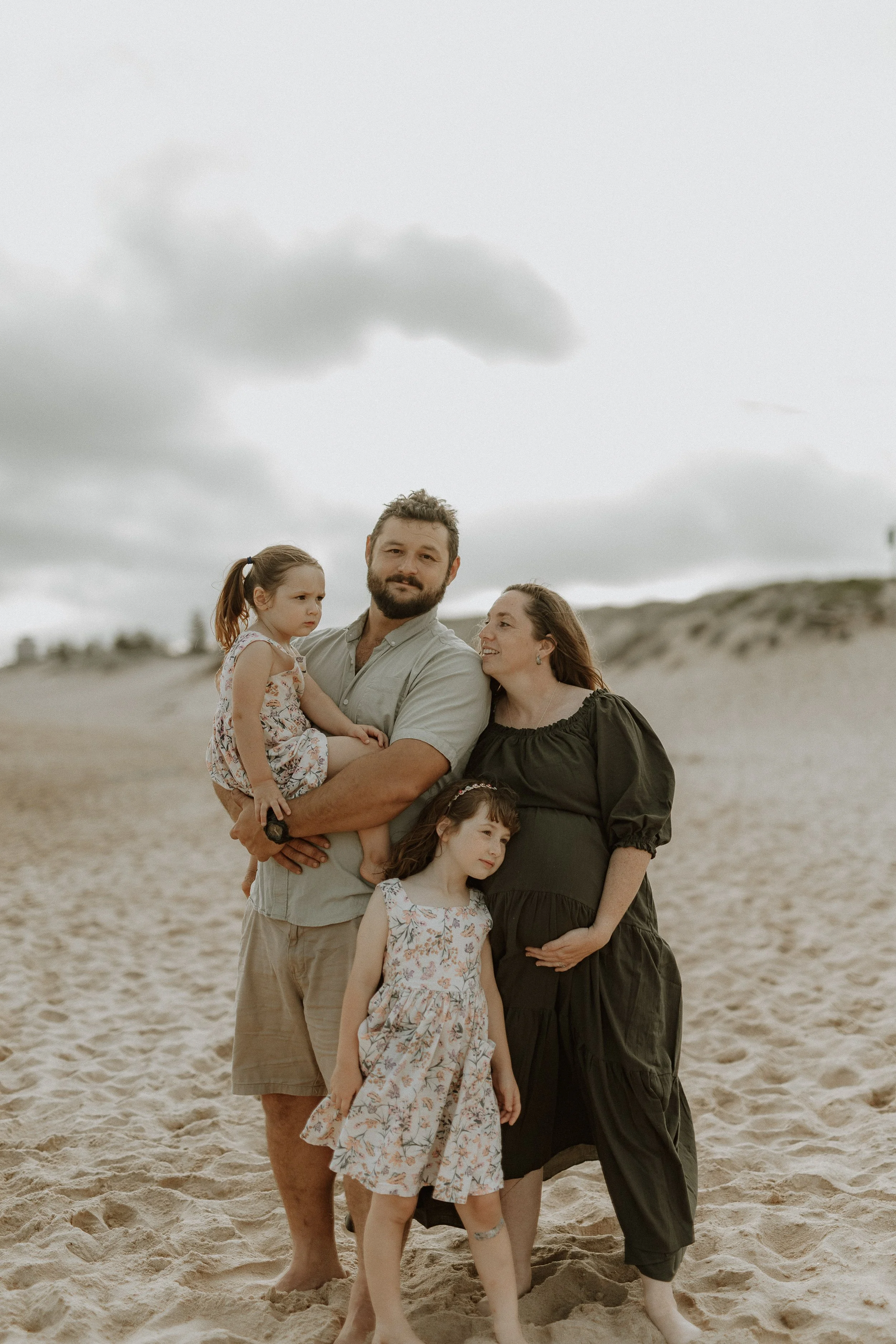 A family of five posing on a sandy beach under a cloudy sky. The man is holding a young girl, and there is a woman and two young girls standing nearby.