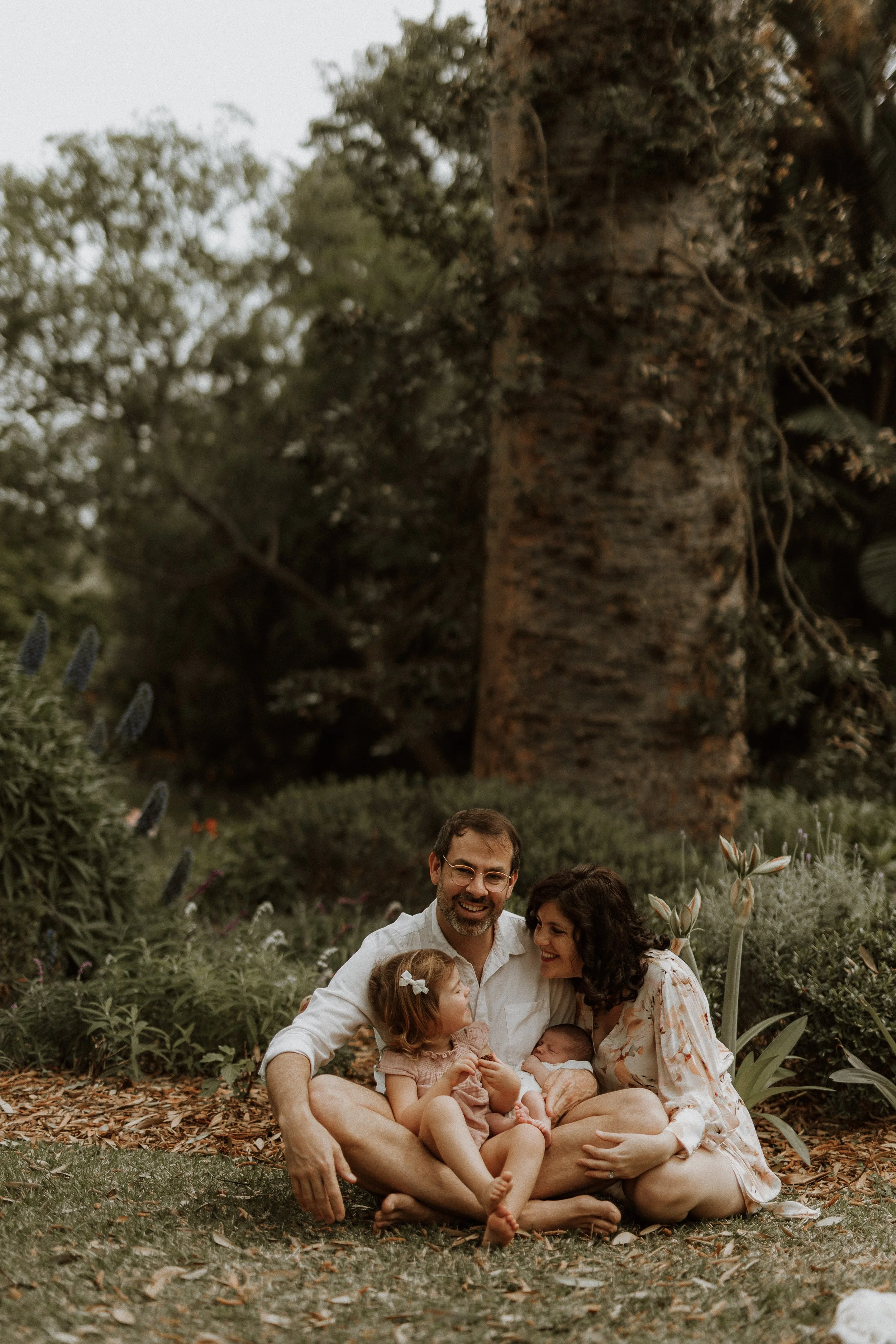 A family of five, including two adults and three children, sitting on the ground in a garden surrounded by flowers and plants, with a large tree in the background, during daytime.