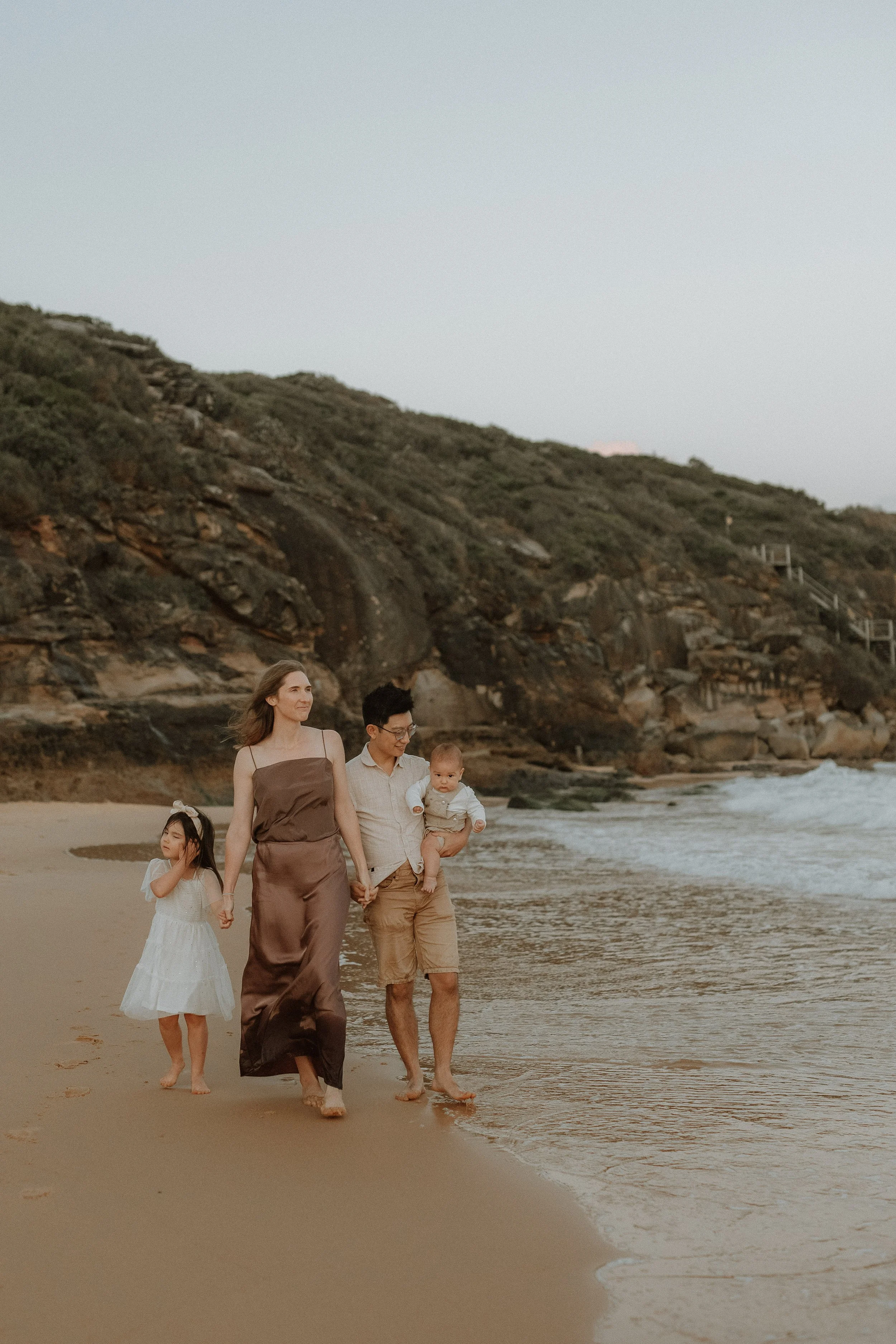 A family walking along the beach during sunset, with rocky hills in the background.