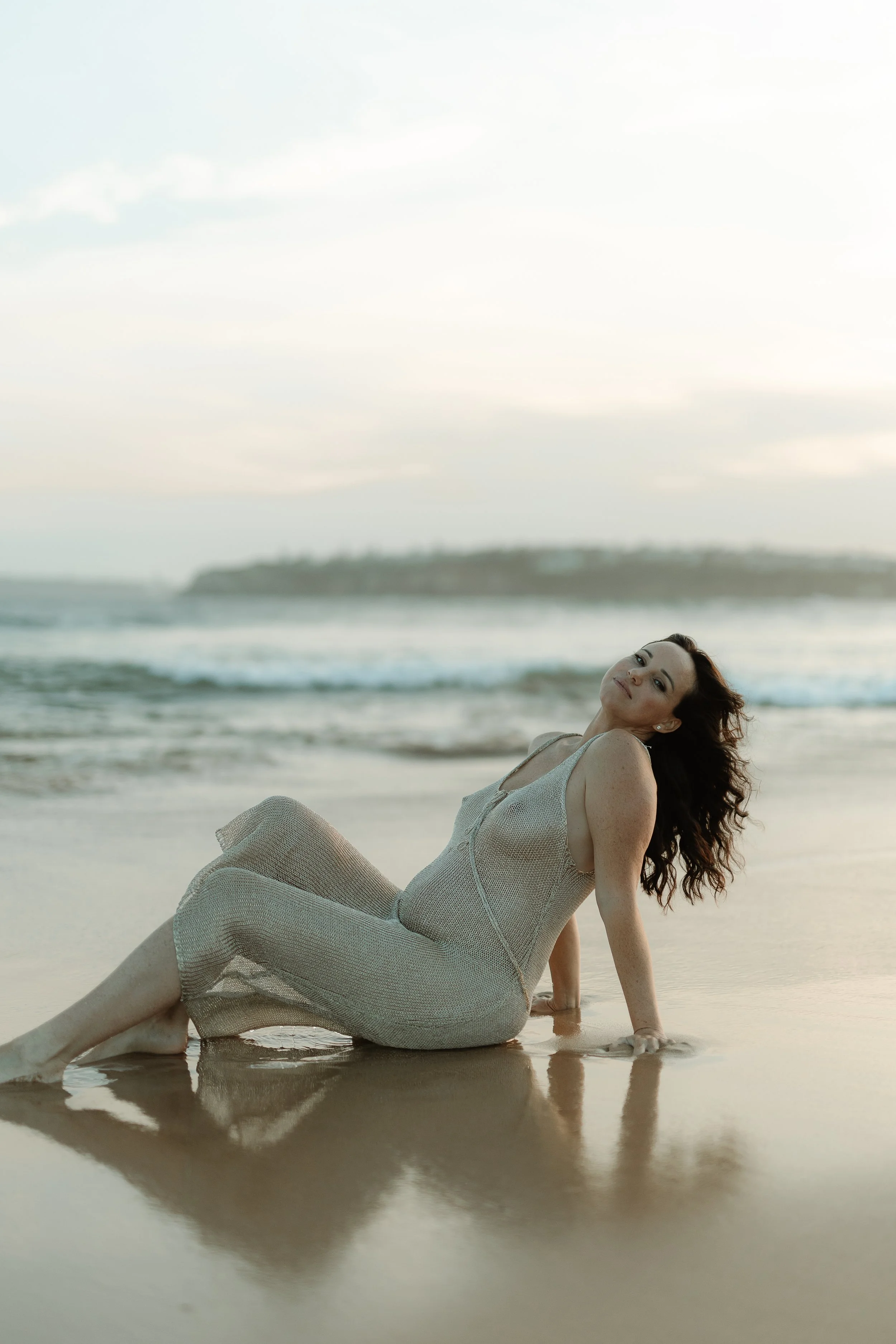 A woman with dark wavy hair lying on a beach at sunset, wearing a light-colored dress, with the ocean and a distant landmass in the background.