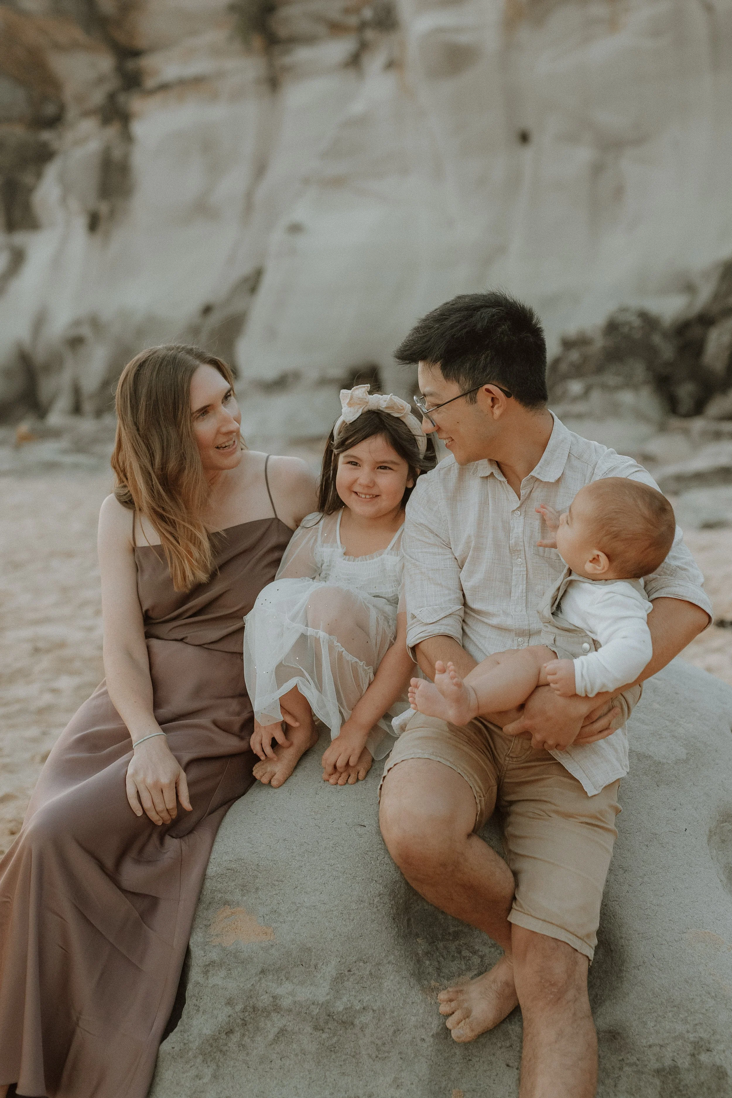 A family of five sitting on a beach, smiling and interacting with each other. The family consists of a woman, a man holding a young child, and two young girls, all appear happy and engaged.