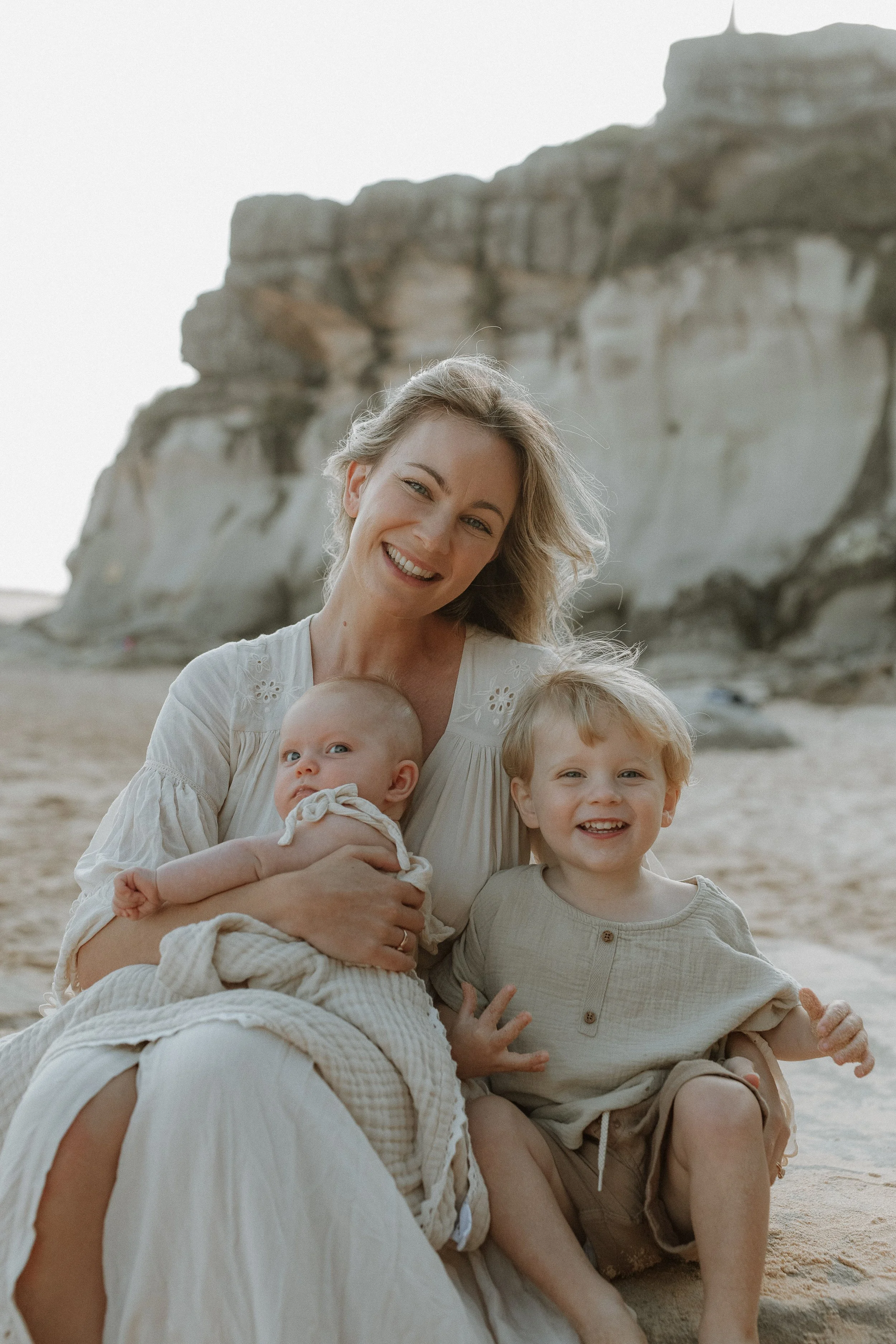 A smiling woman with curly blonde hair holding a baby and sitting next to two young boys at the beach with rocky cliffs in the background.