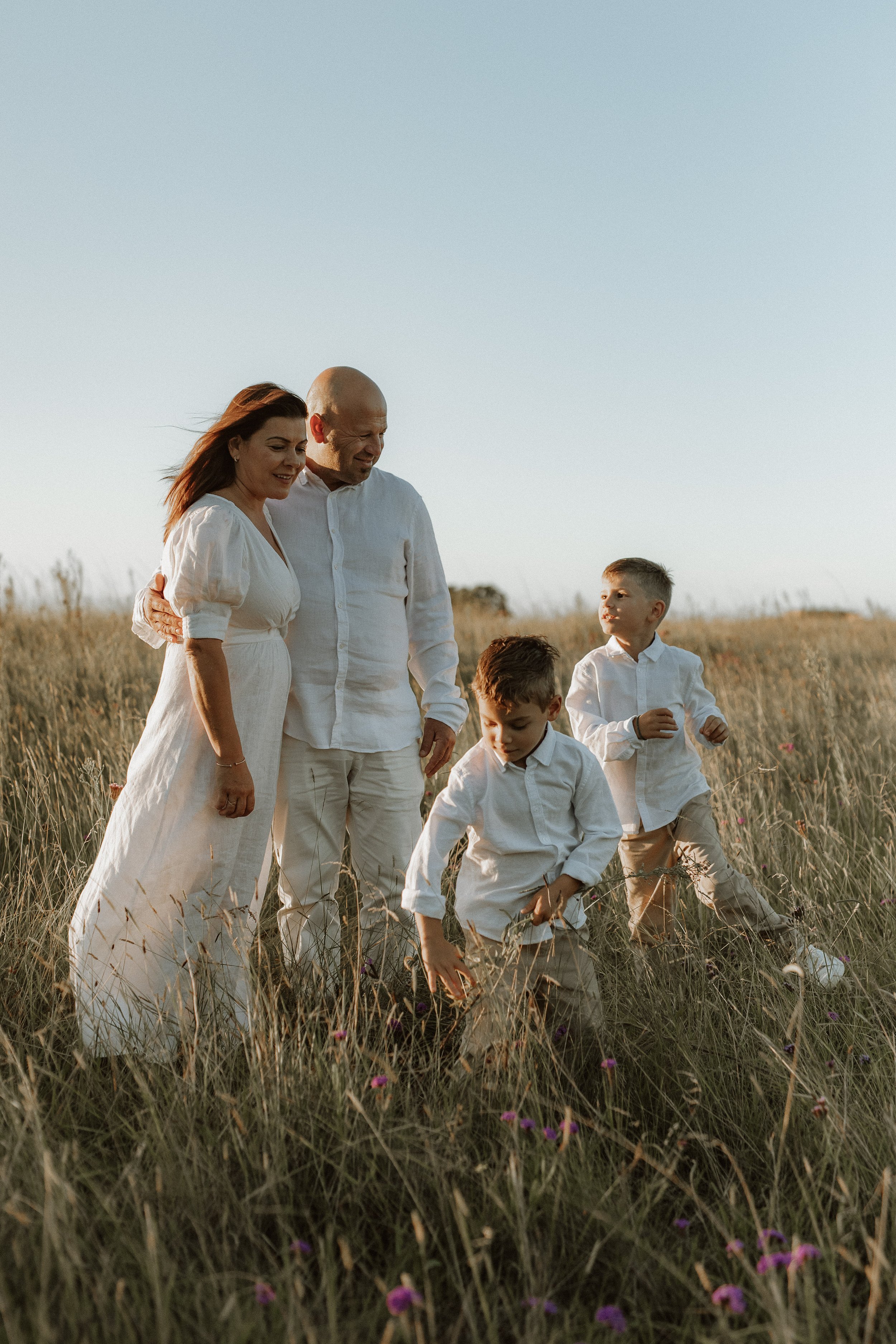 A family of five, including a woman, two men, and two boys, standing in a field of tall grass and small purple flowers, all wearing light-colored clothing, enjoying a sunny day.