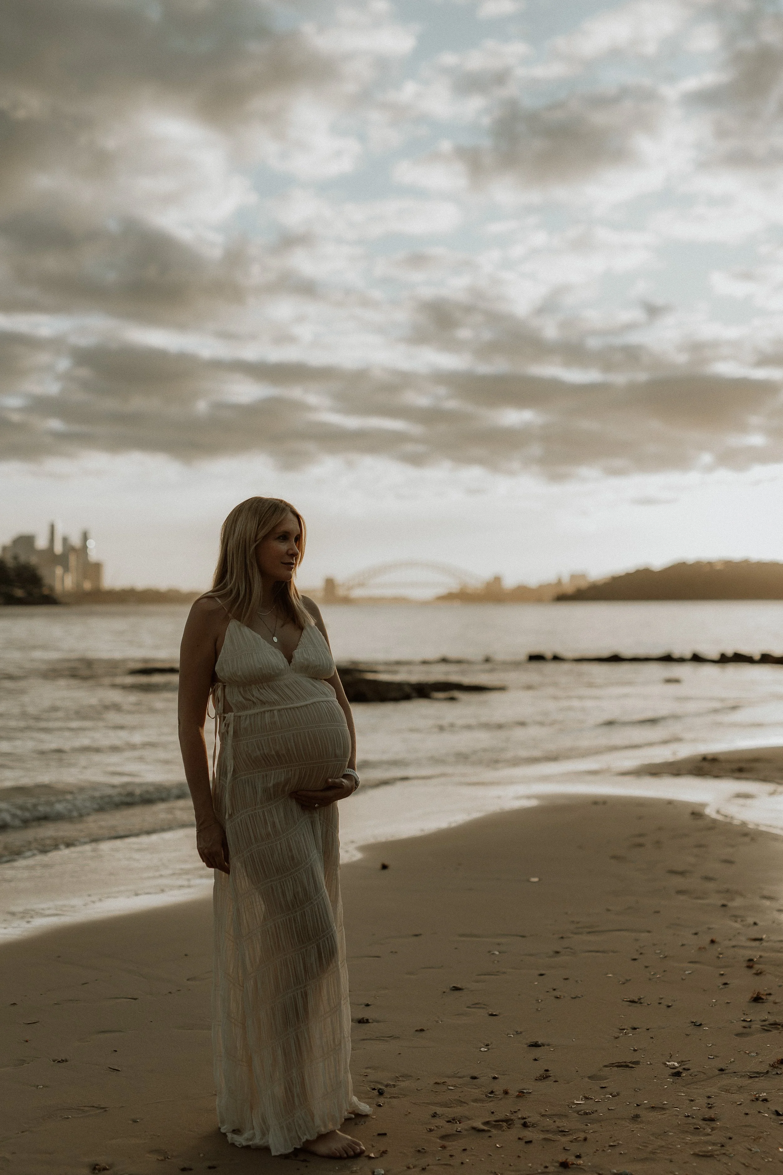 Pregnant woman standing barefoot on the beach during sunset, wearing a long beige dress, with city skyline and bridge in the background under cloudy skies.