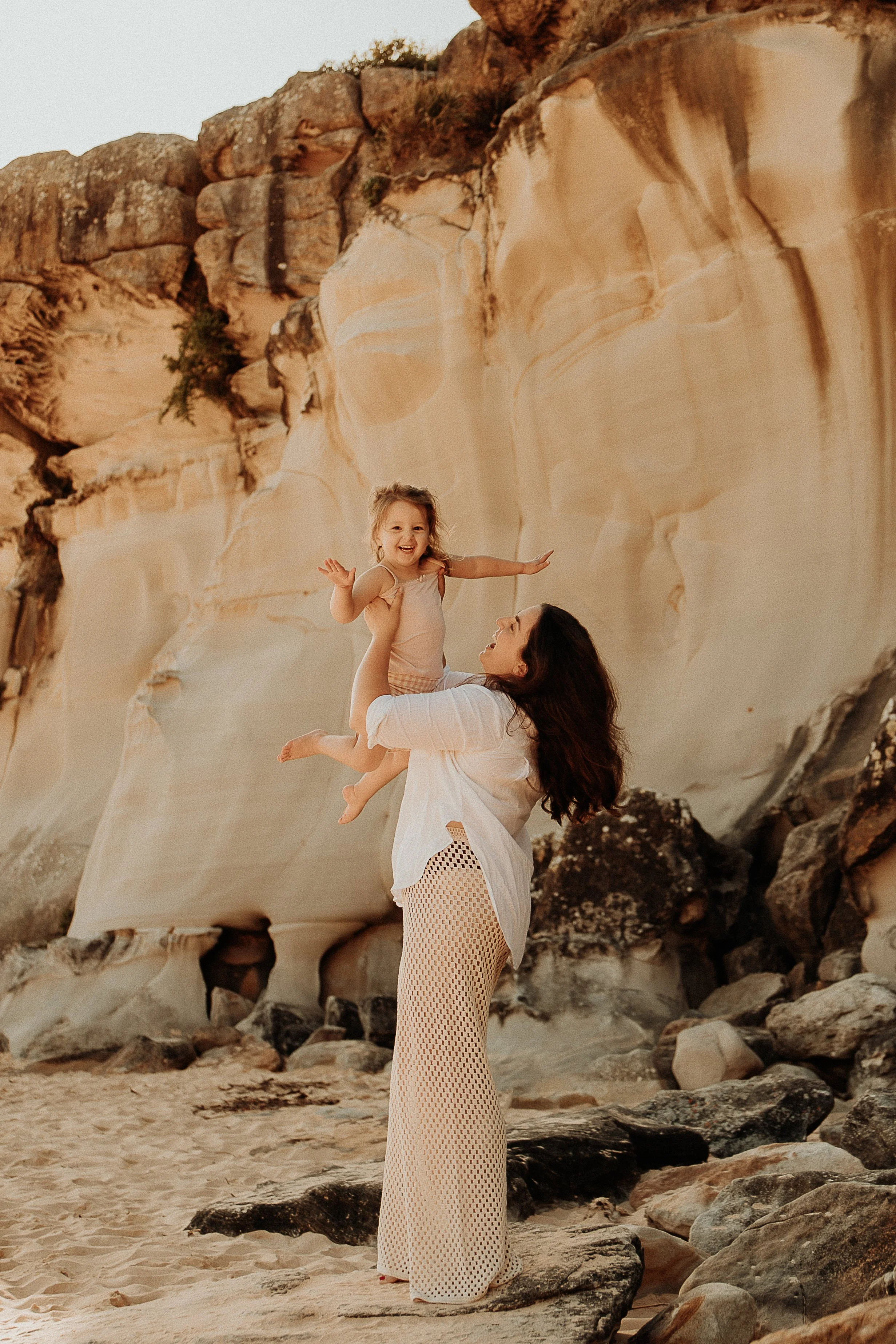 Woman holding a child in front of a rocky cliff on a sandy beach.