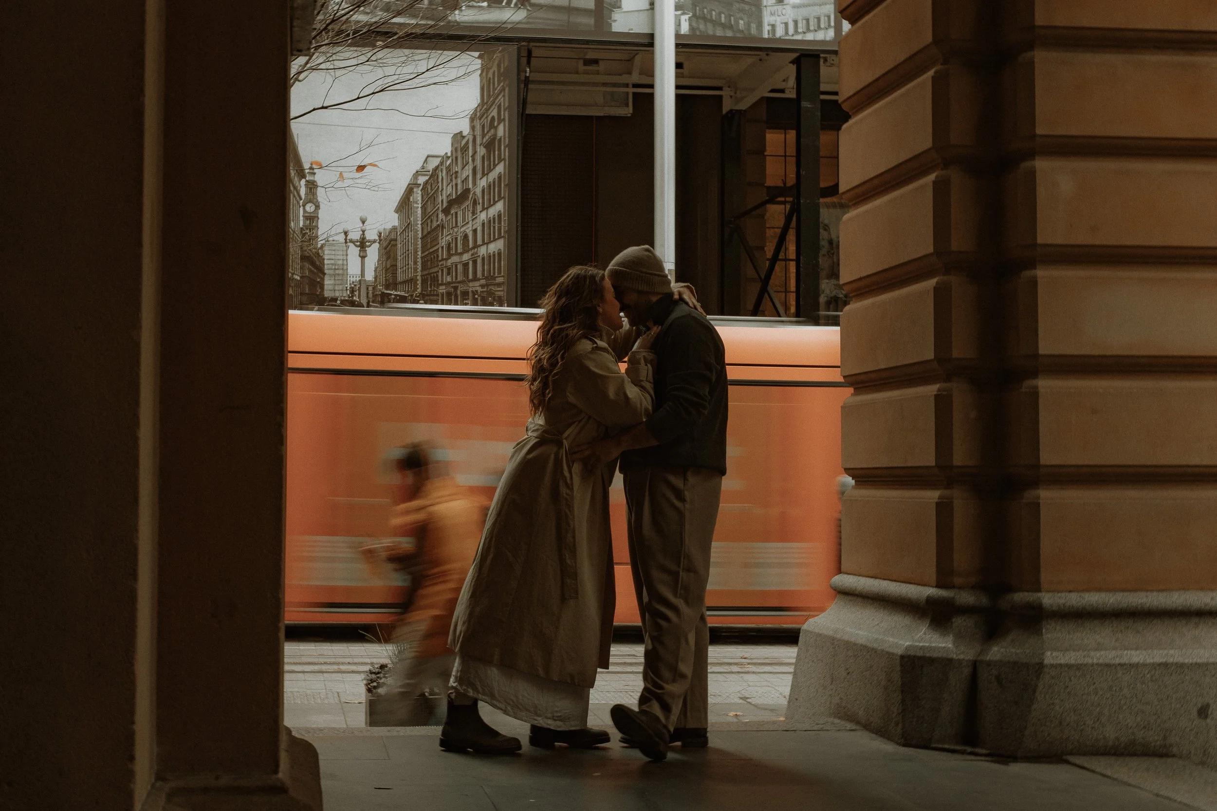 A couple standing close and embracing while about to kiss, seen through an opening between buildings with a city street in the background