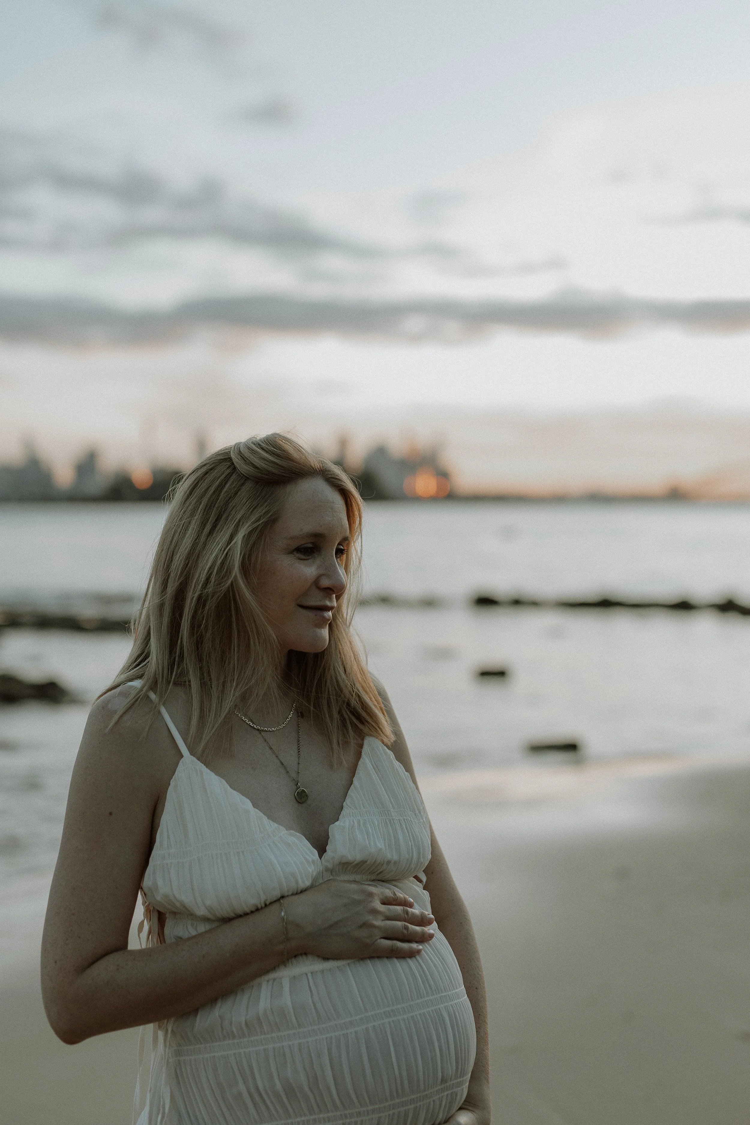 A pregnant woman with long blonde hair wearing a white dress, standing on a beach at sunset, with her hand resting on her belly and a city skyline visible in the background.