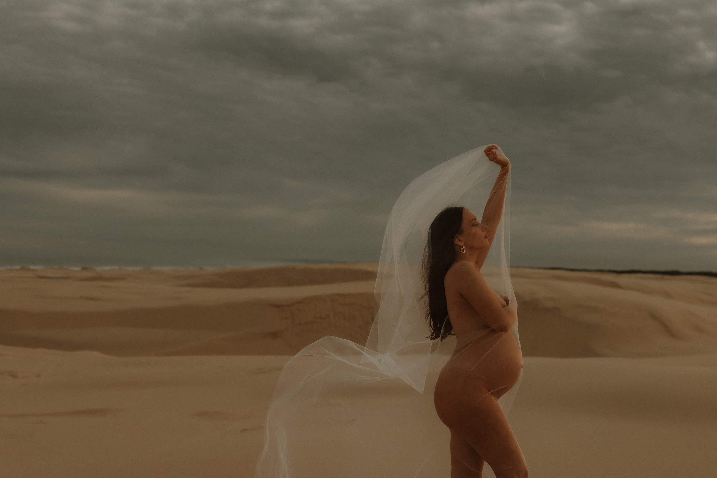 A woman with long dark hair standing nude on a sandy beach under a cloudy sky, holding a sheer white fabric above her head.