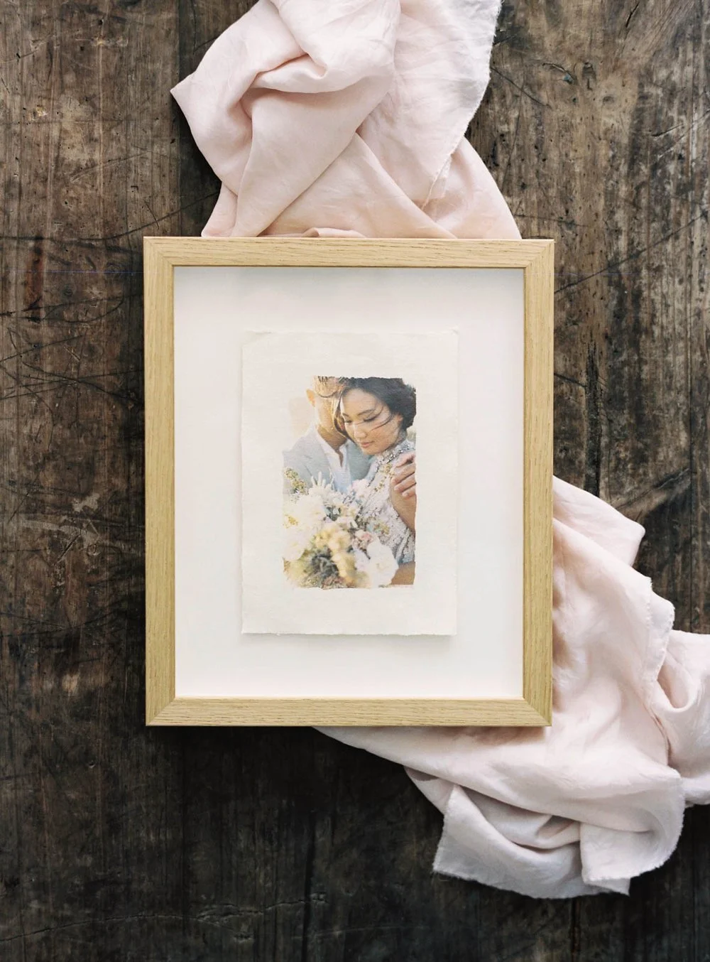 Framed photo with a light wood frame on a textured surface, featuring a couple embracing with a bouquet, and surrounded by a soft pink fabric.