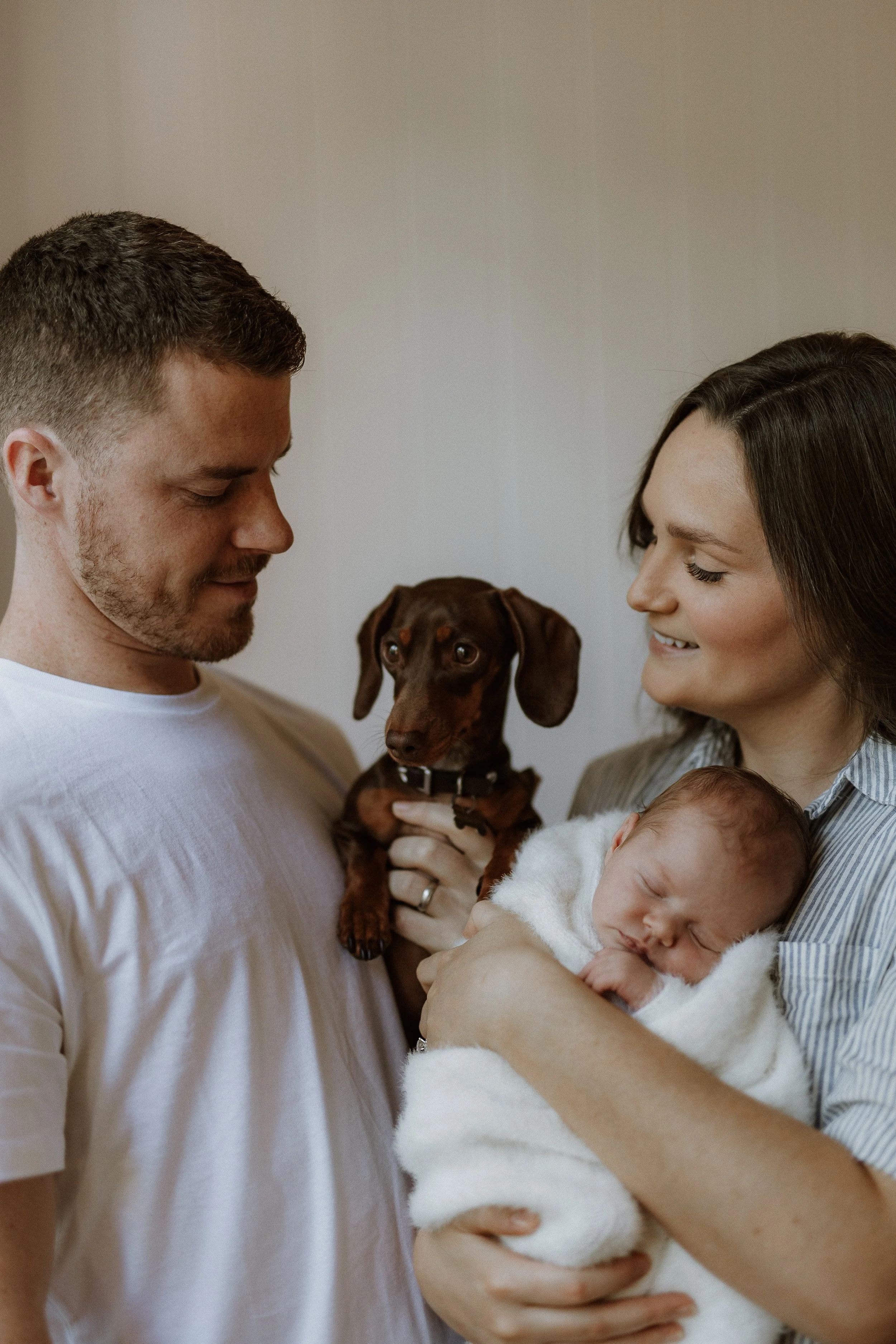A man and woman smiling at each other, while holding a baby and a dog.