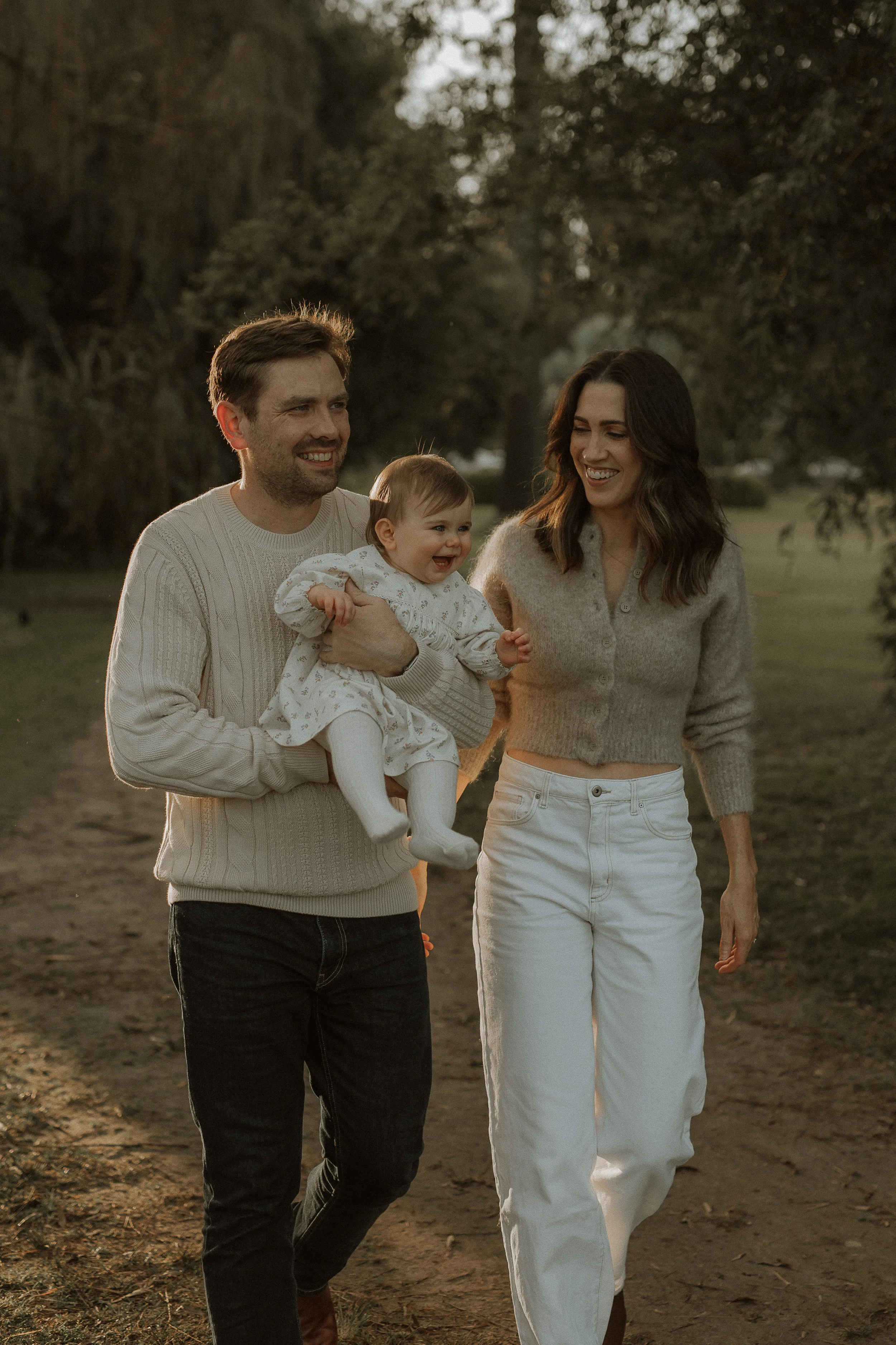 A family of three walking outdoors at sunset. The man is holding a smiling toddler girl, and a woman is walking beside them.