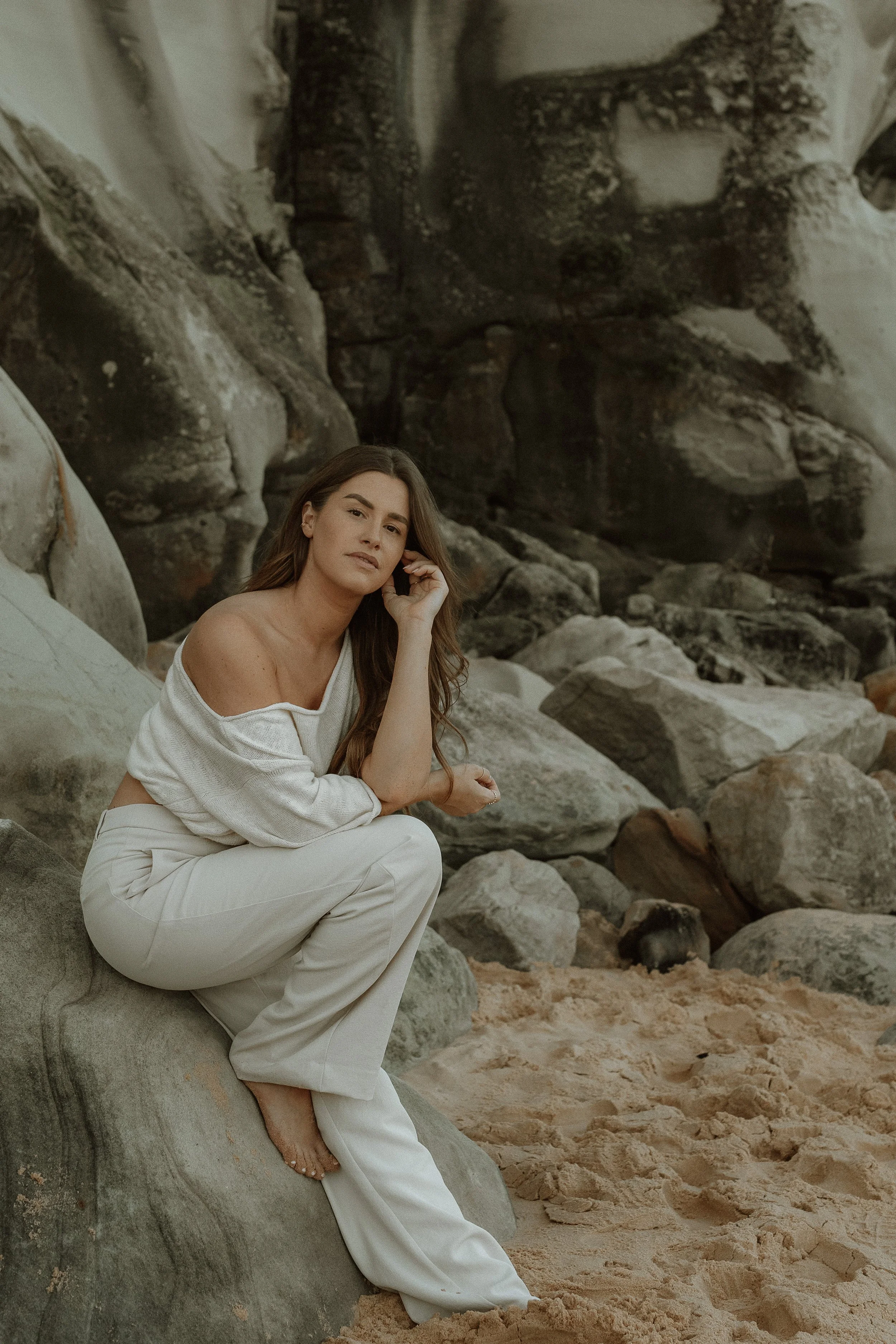 A woman sits on a large rock formation on a beach with sand, wearing a white off-shoulder top and white pants, with long brown hair, in a relaxed pose.