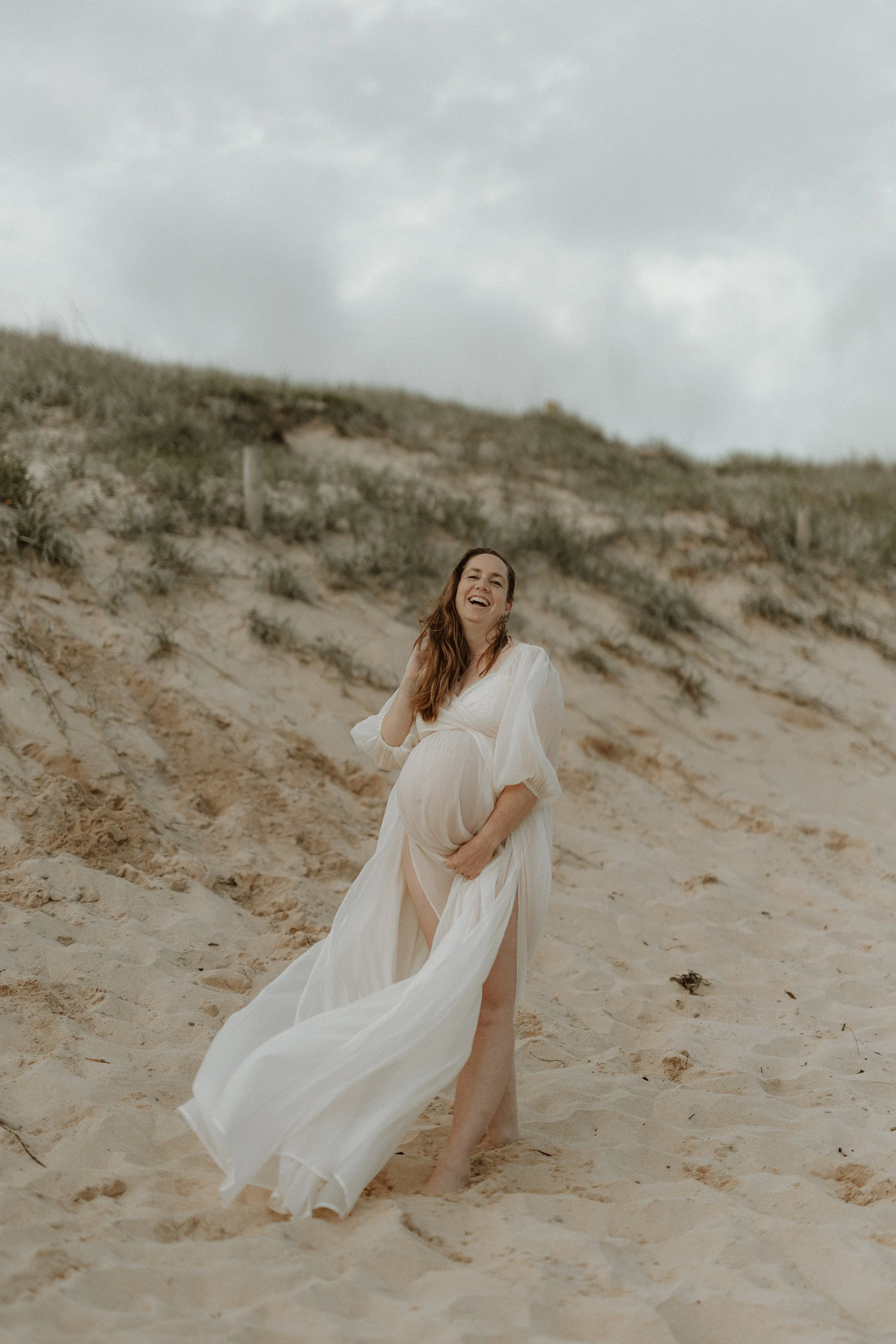 Woman in a white dress standing barefoot on a sandy beach, smiling, with a dune and cloudy sky in the background.