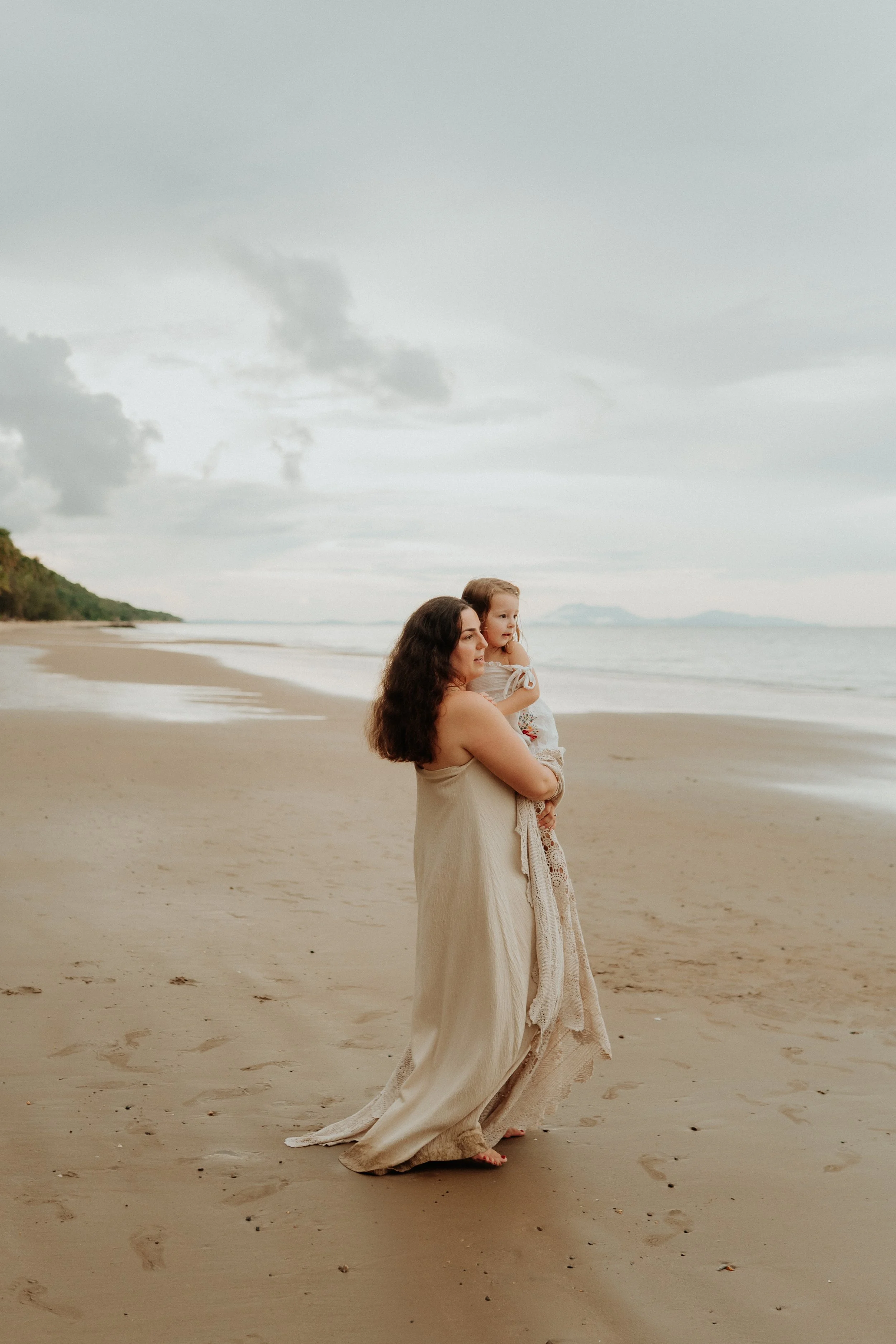 A woman holding a young girl standing on a cloudy beach with footprints in the sand and distant landforms on the horizon.