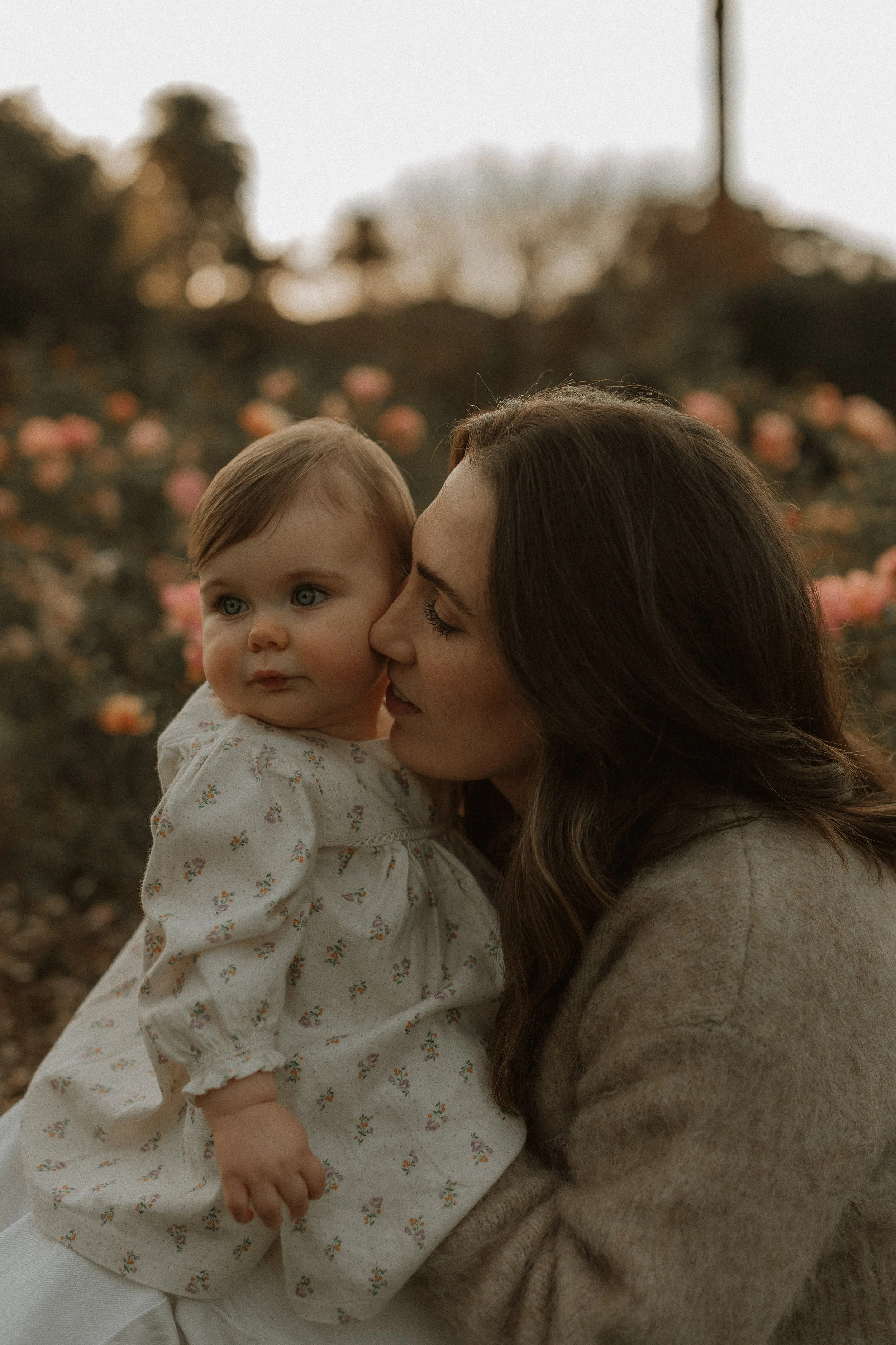 A woman with long brown hair holding a young girl with light brown hair and blue eyes, outdoors during a sunset with trees and pink flowers in the background.