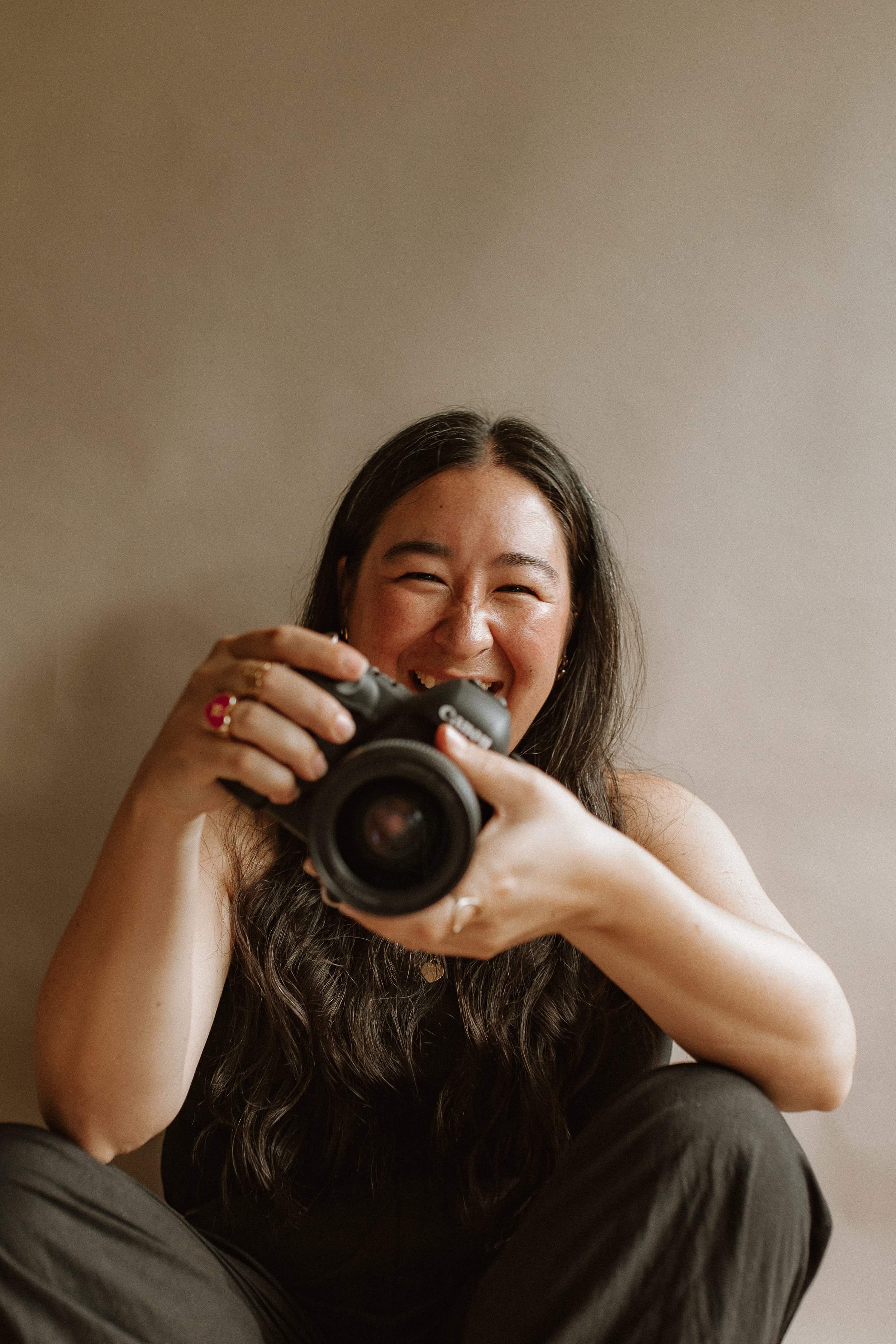 A woman with long dark hair taking a selfie with a camera and smiling, sitting against a plain background.